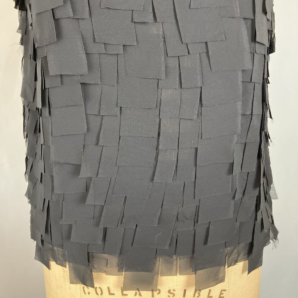A close-up of a dark gray fabric accessory, likely a skirt or wrap, covered in layered rectangular fabric strips. The bottom edge shows a beige mannequin or display form with the word 'COLLAPSIBLE' partially visible.