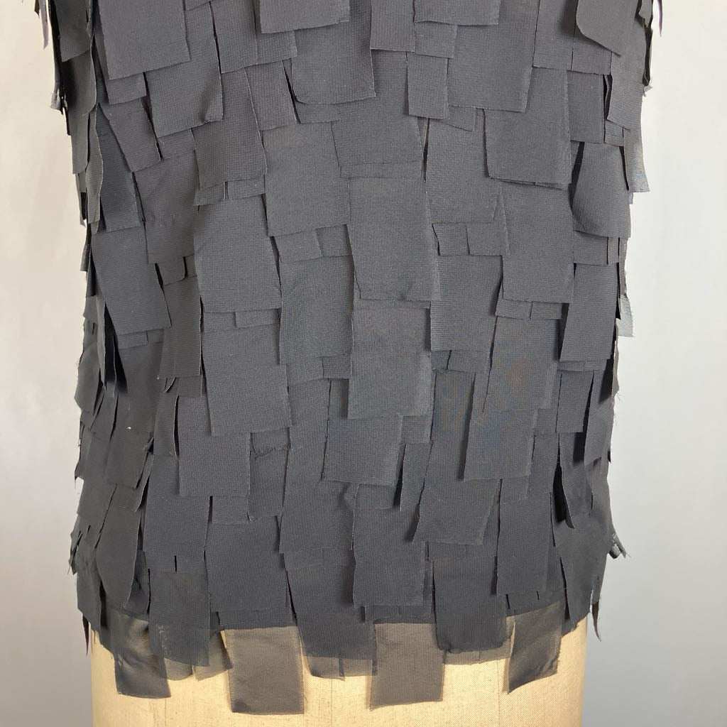 A dark gray fringed tunic top with layered rectangular fabric pieces, displayed on a beige dress form against a light gray background.