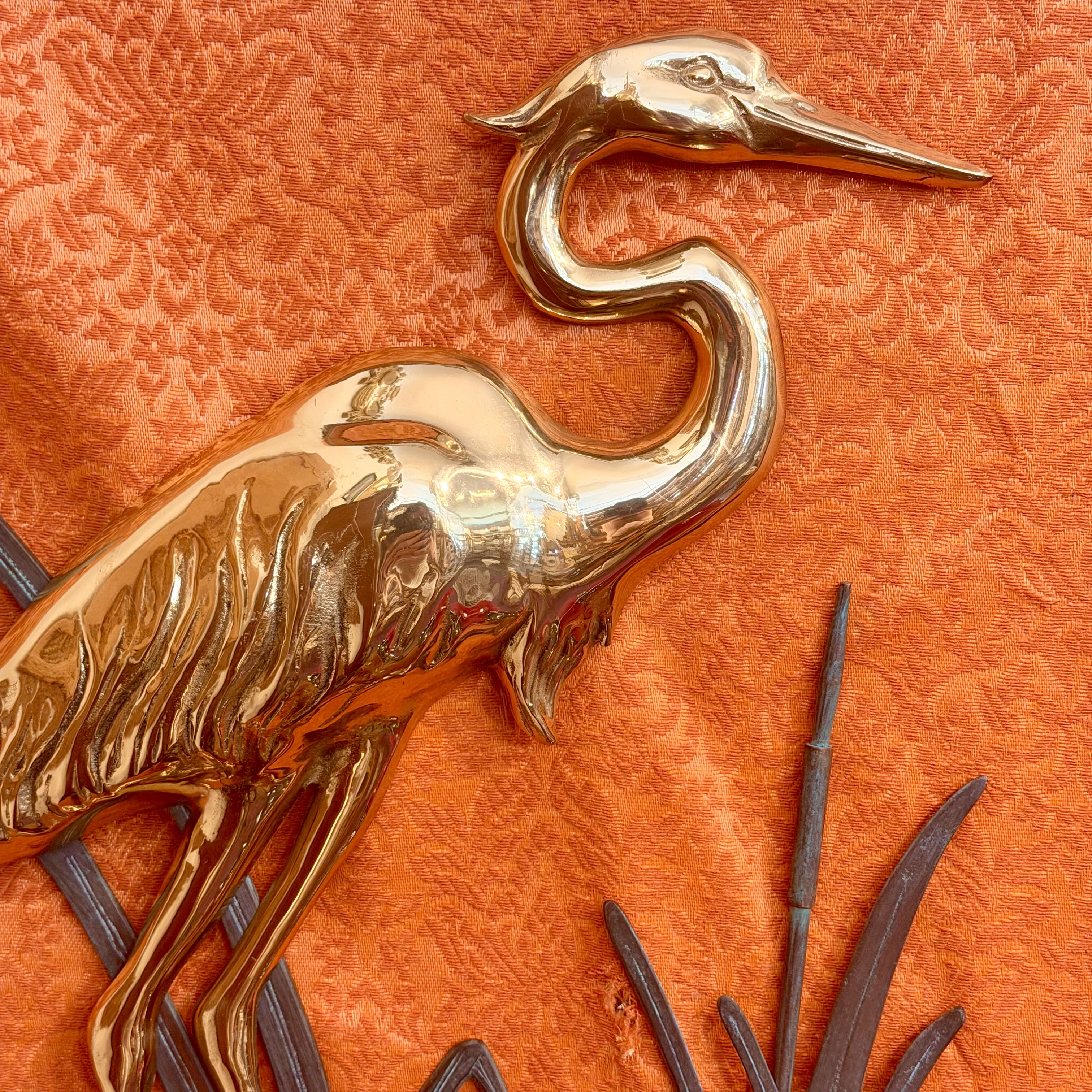 A polished gold-toned metal heron wall hanging mounted on an orange damask fabric background.