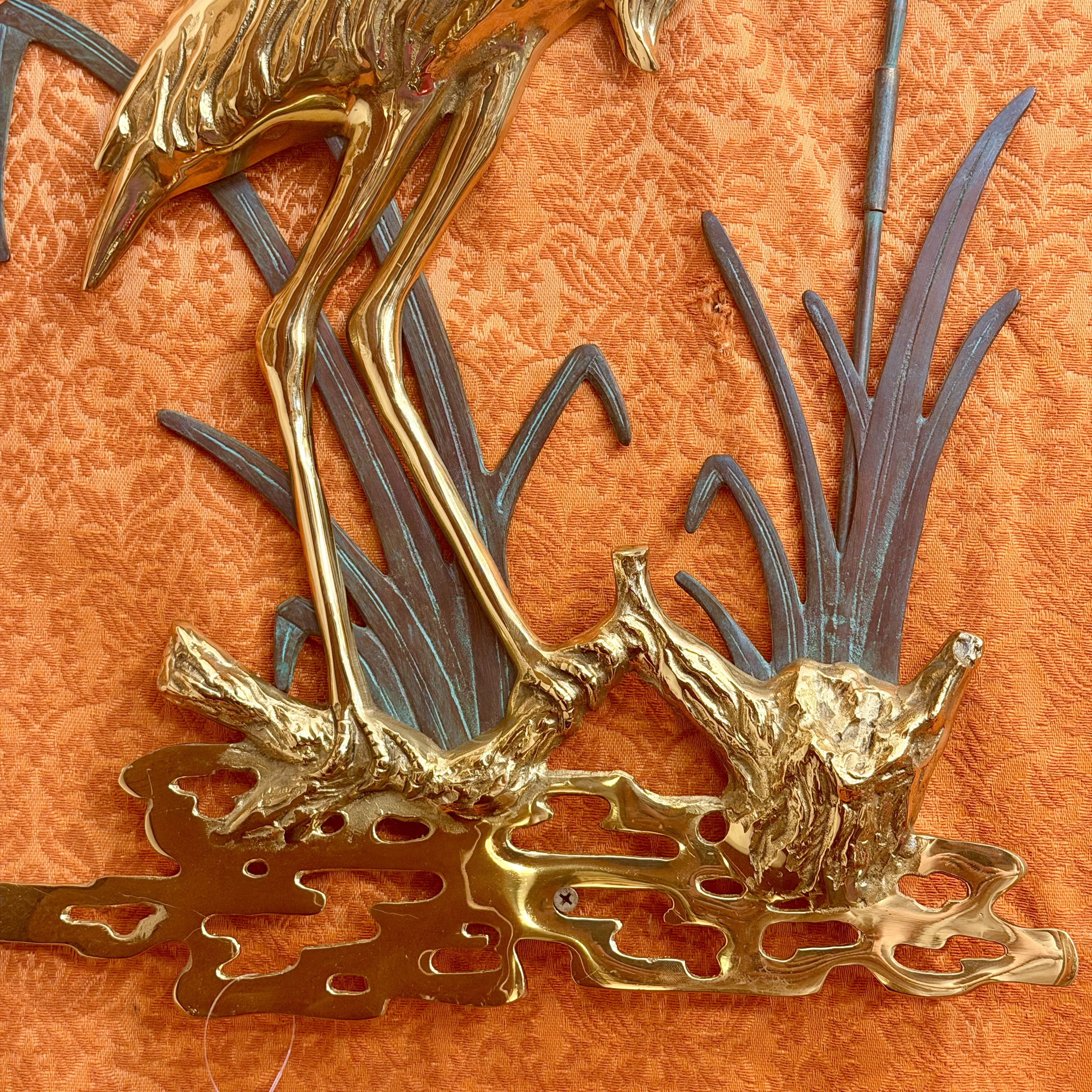 A close-up of a decorative wall sculpture featuring a golden bird standing among stylized reeds and branches on a carved base, set against an orange patterned fabric background.