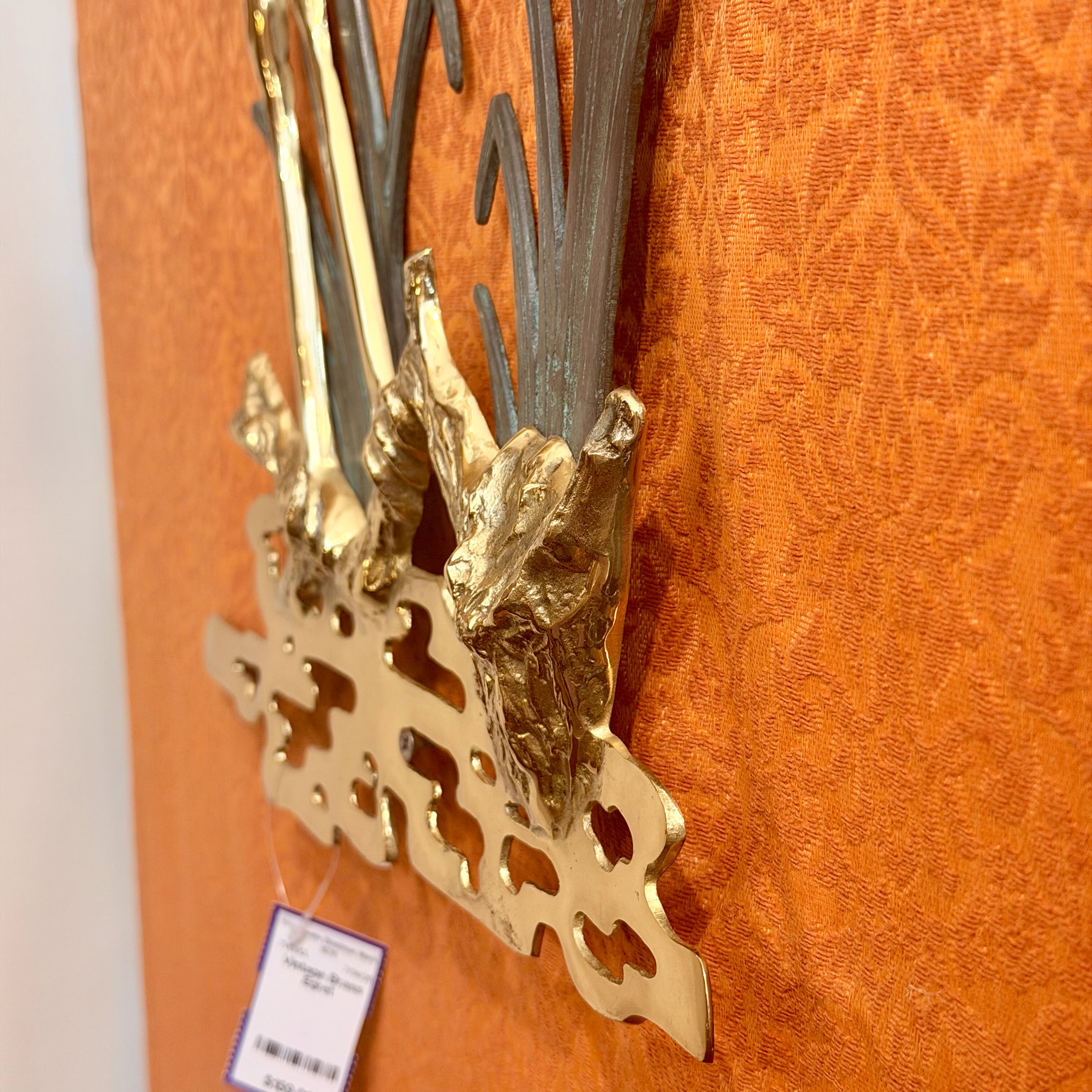 A close-up of a decorative wall-mounted metal piece with ornate gold-toned and dark gray branches or leaf-like elements, attached against an orange patterned fabric backdrop. A small product tag hangs from one end.
