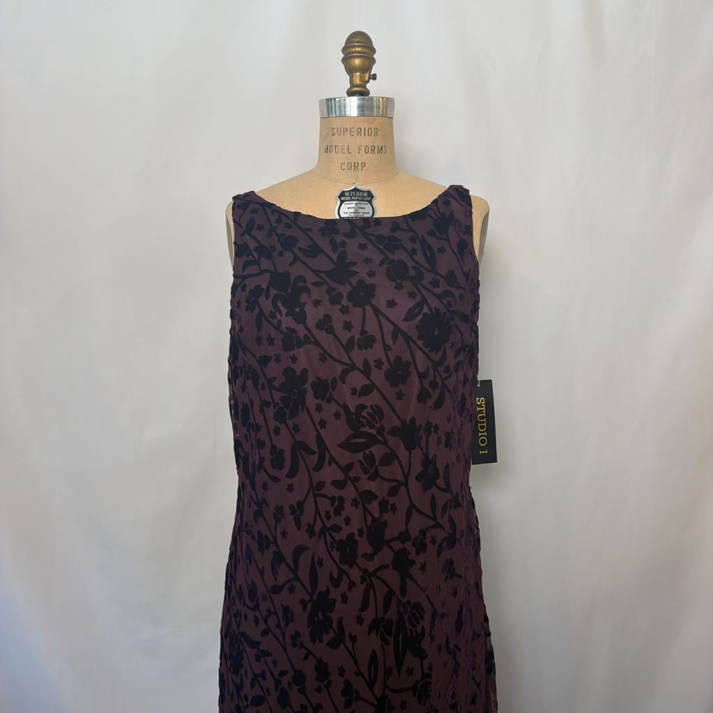 A sleeveless dark plum dress with a black floral pattern, displayed on a dress form mannequin against a white fabric backdrop. A black 'STUDIO' brand tag is visible on the side.