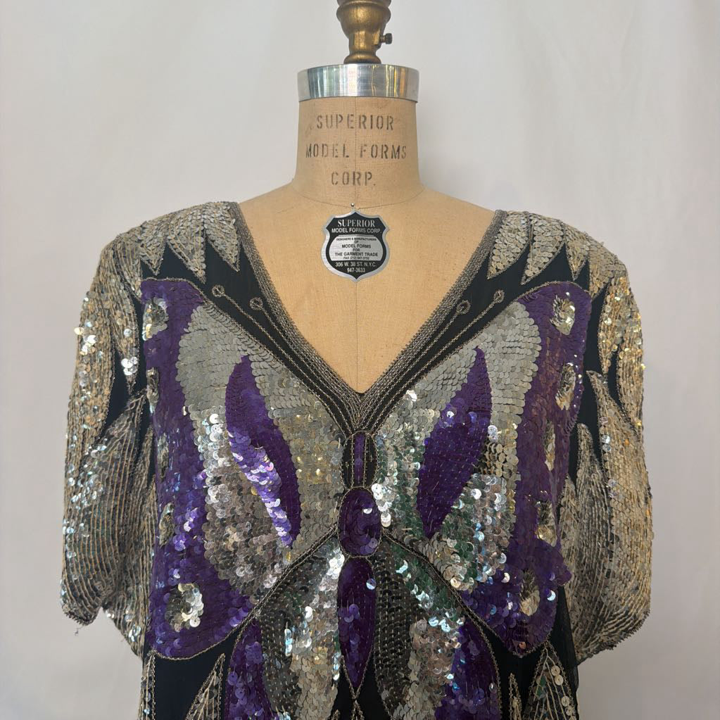 A vintage sequined top displayed on a tan dress form mannequin. The top features a V-neckline, short puff sleeves, and a butterfly-wing motif in purple, silver, and black sequins.