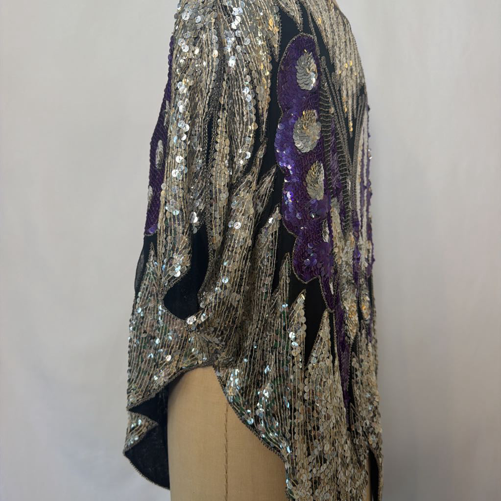 A close-up of a sequined garment draped on a mannequin torso, featuring metallic silver and gold sequins with a prominent purple sequined applique detail on a light gray background.