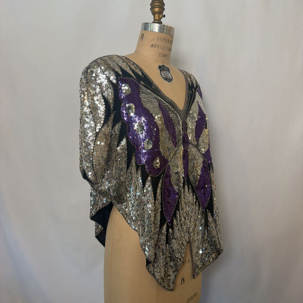 A vintage-style sequined blouse displayed on a dress form. The garment features a silver sequin base with a large purple sequined butterfly motif on the front, a V-neckline, and short draped sleeves.