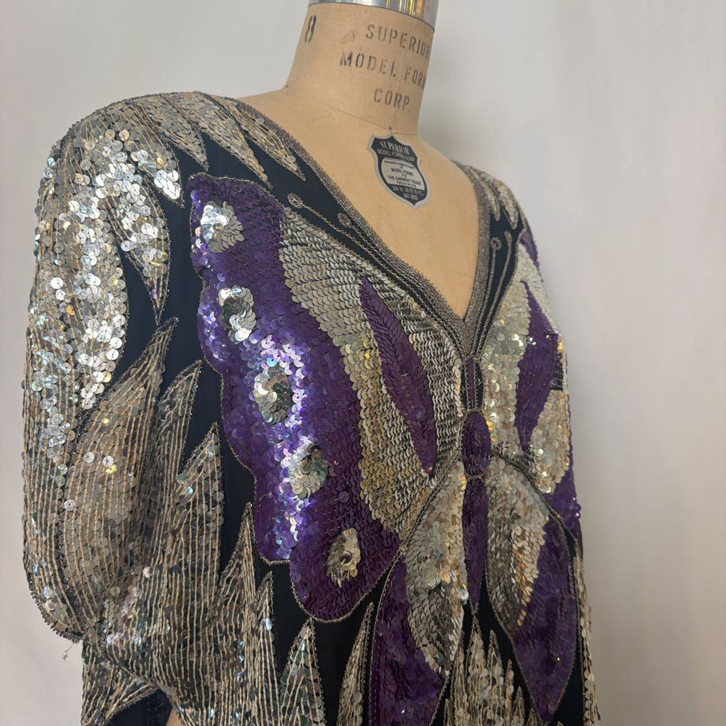 A close-up of a vintage 1920s flapper-style beaded blouse displayed on a dress form. The garment features intricate silver and gold sequins with prominent purple velvet butterfly wings on a black base.