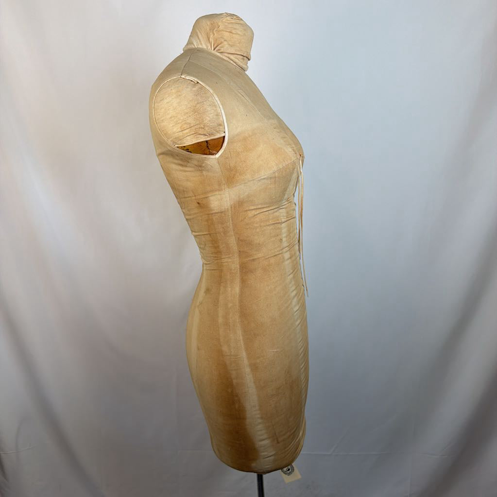 A beige satin or silk dress form mannequin torso with a shoulder cutout, standing on a black stand against a white fabric backdrop.