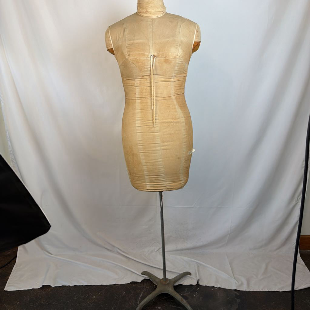 A beige textured dress form mannequin torso with a front zipper, mounted on a metal stand against a draped white fabric backdrop.