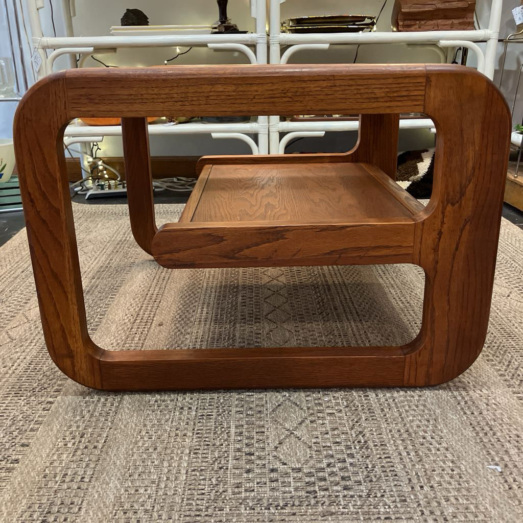 A mid-century modern wooden coffee table with a warm brown finish, featuring a thick rounded rectangular frame and a lower shelf, set on a textured beige rug.