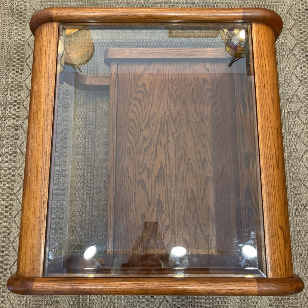 A square wooden coffee table with a clear glass front and rounded corners, featuring a medium brown wood grain finish and built-in lighting.