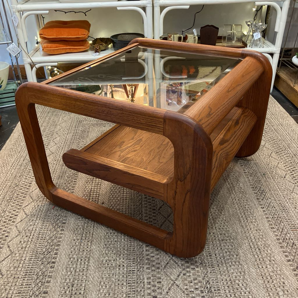 A mid-century modern wooden coffee table with a glass top, rounded rectangular frame, and open lower shelf, shown on a textured rug in a shop setting.