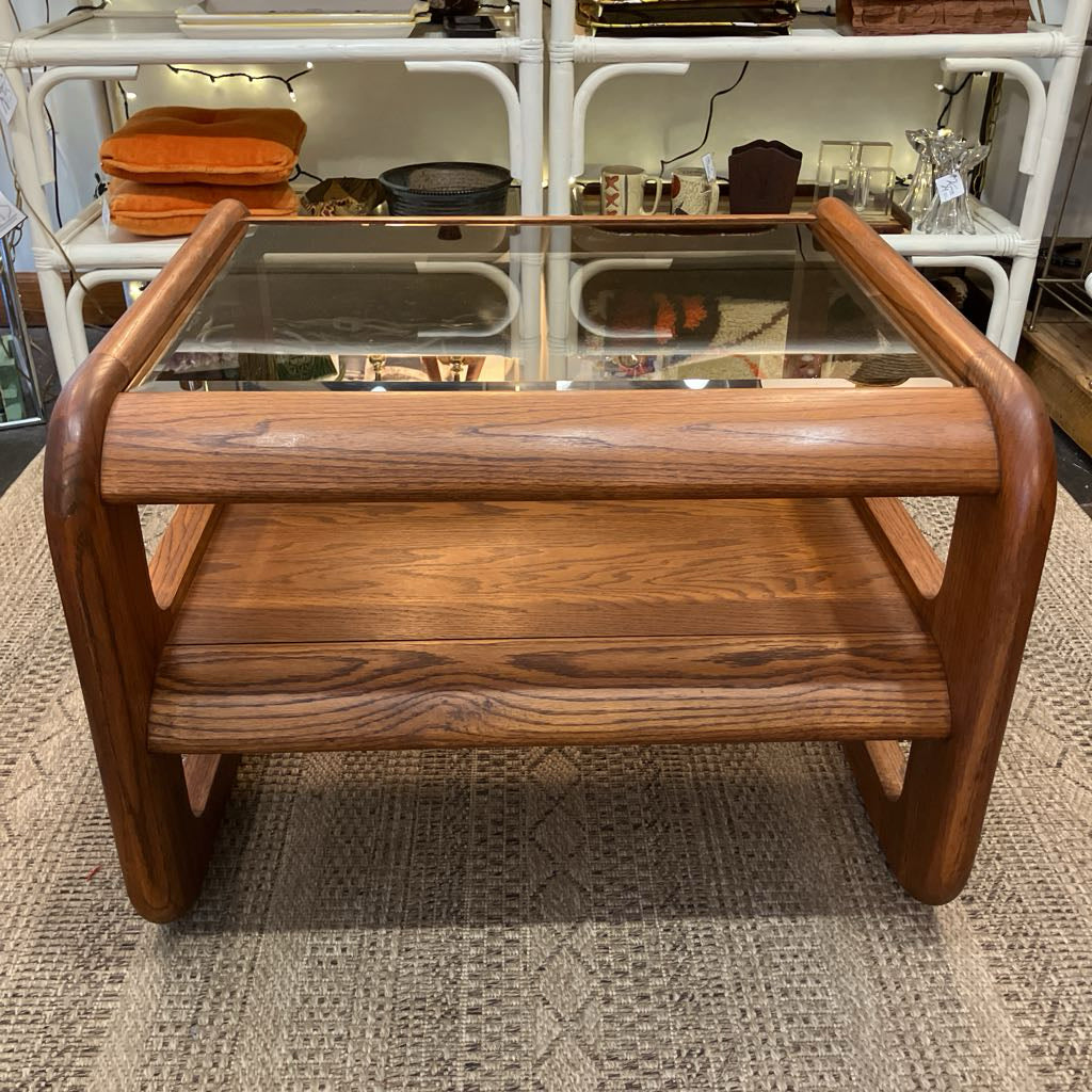 A mid-century modern wooden coffee table with a glass top, two wooden shelves, and curved legs, set on a textured rug in front of white metal shelving.