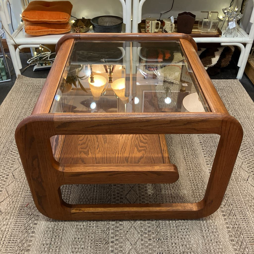 A mid-century modern wooden coffee table with a thick warm brown wood frame, rounded corners, and a clear glass top, set on a textured beige rug in a showroom.