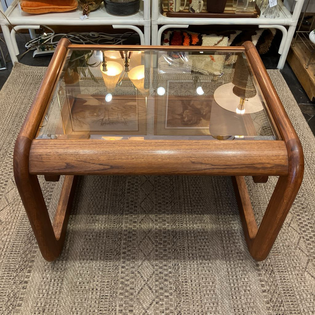A mid-century modern wooden coffee table with a clear glass top and curved walnut-look wood legs and apron, shown on a patterned rug in a furniture store.