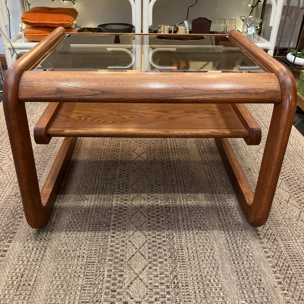 A mid-century modern wooden coffee table with a glass top, a lower shelf, and curved legs, shown on a patterned rug in what appears to be a furniture showroom.
