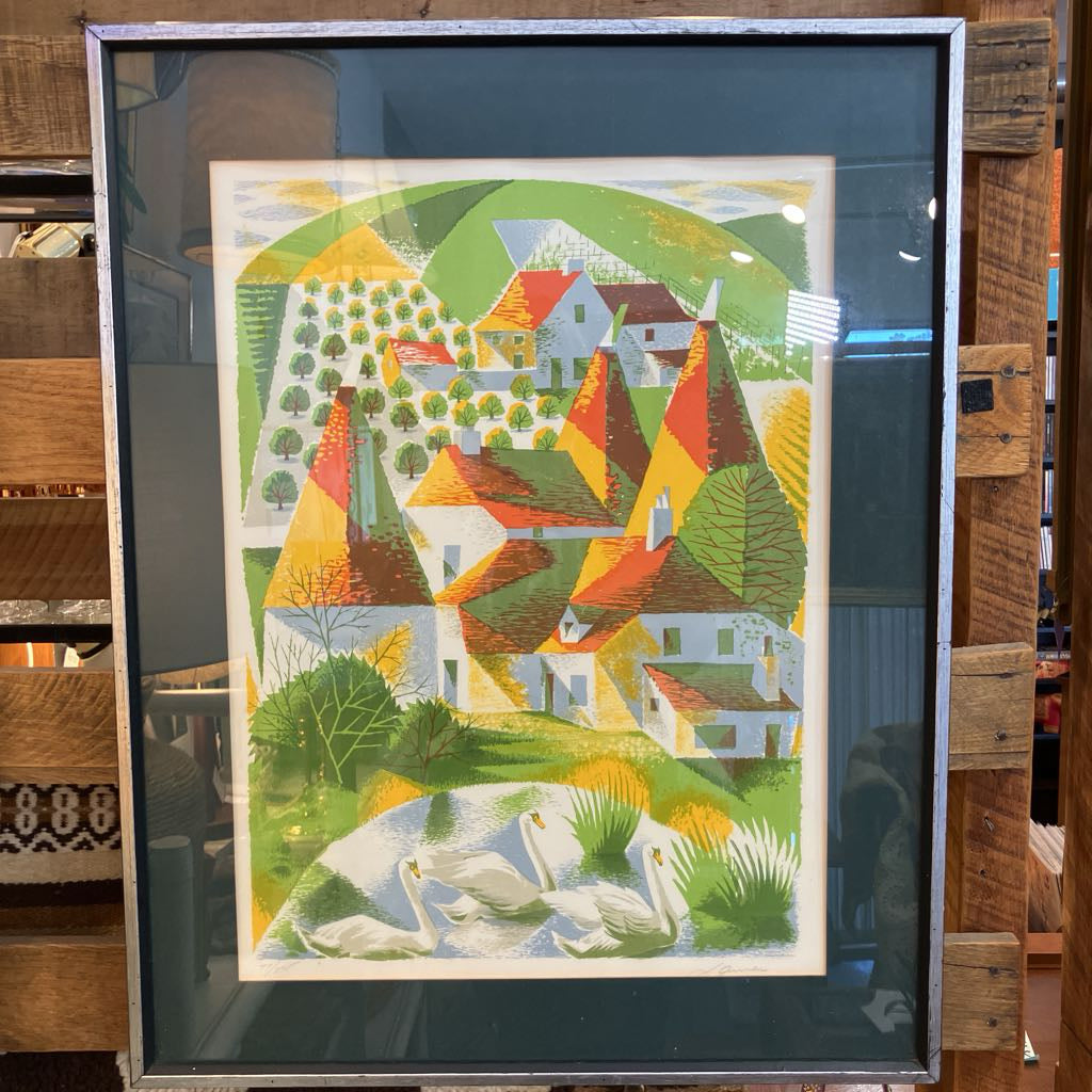A framed art print featuring a colorful stylized landscape with houses, trees, and swans, displayed in a dark frame against a rustic wooden background.