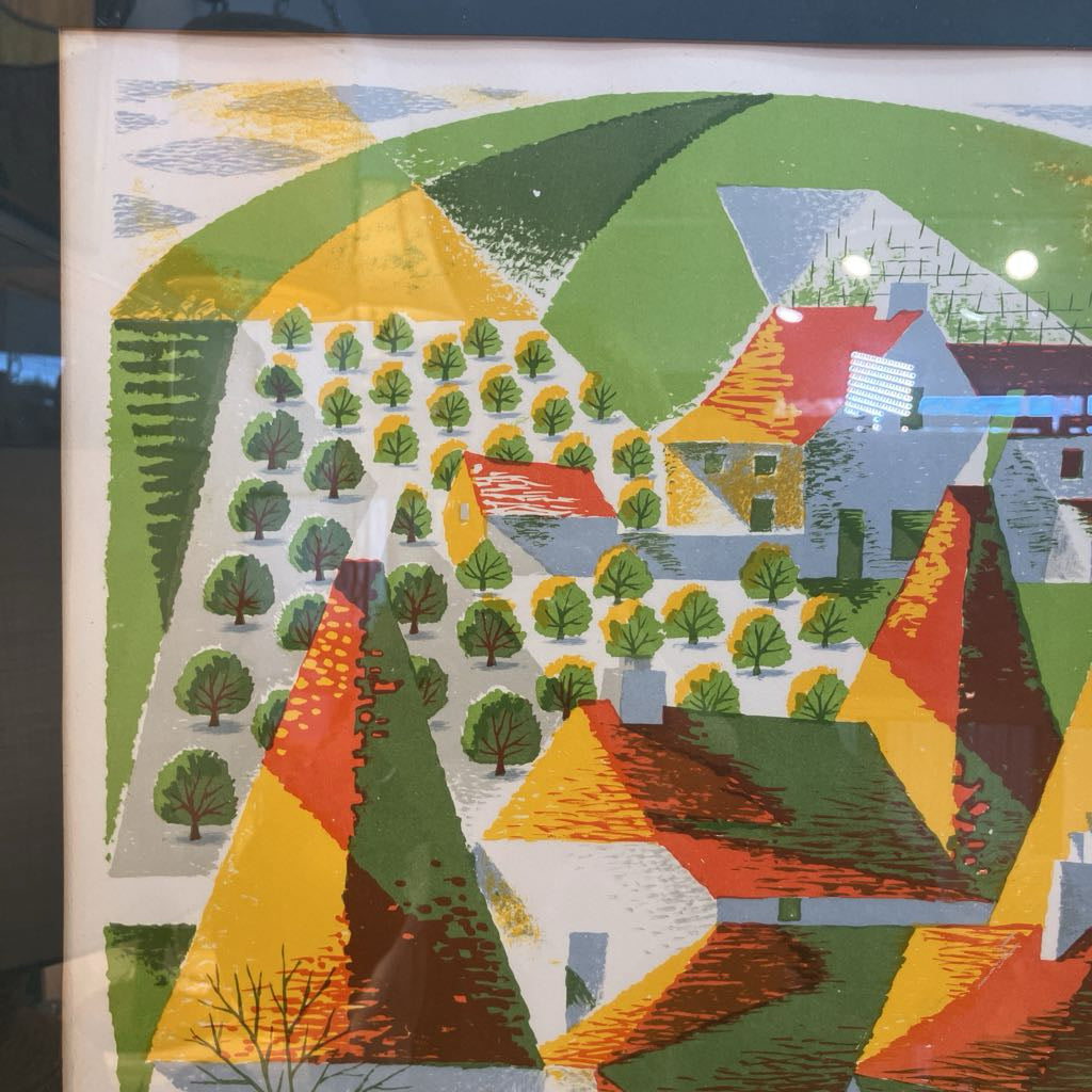 A colorful abstract landscape art print featuring stylized hills, trees, and houses in green, yellow, orange, red, and gray tones, photographed indoors with visible reflections on the glass.