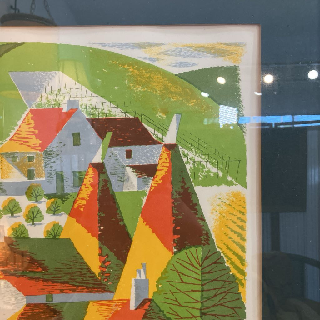 A framed art print depicting a stylized landscape with green hills, yellow fields, and houses with red and brown roofs, shown in a gallery setting with reflections on the glass.