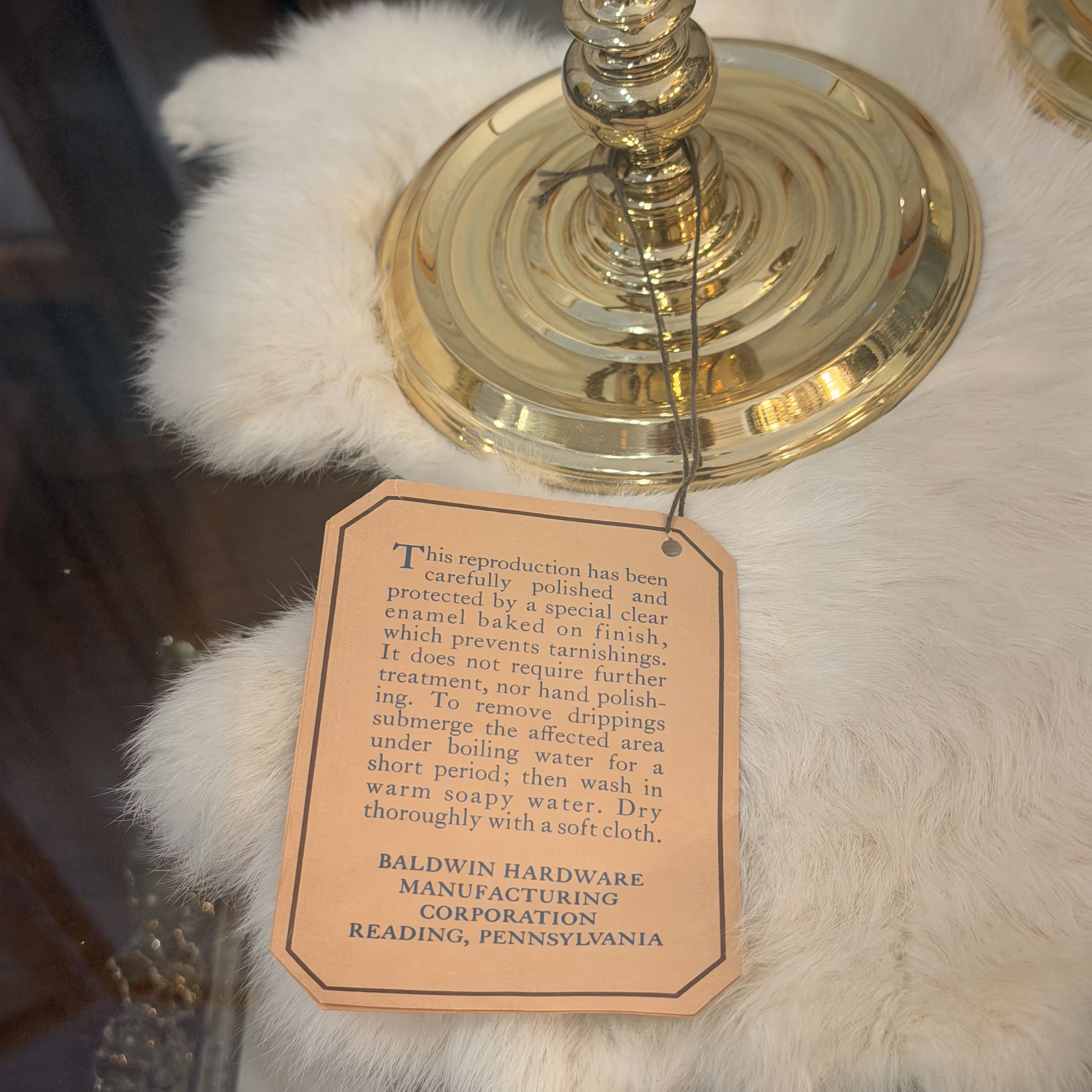 A close-up of a white faux fur or fur textile draped over a gold metal stand, with a hanging tan tag containing care and reproduction information. The tag reads Baldwin Hardware Manufacturing Corporation.