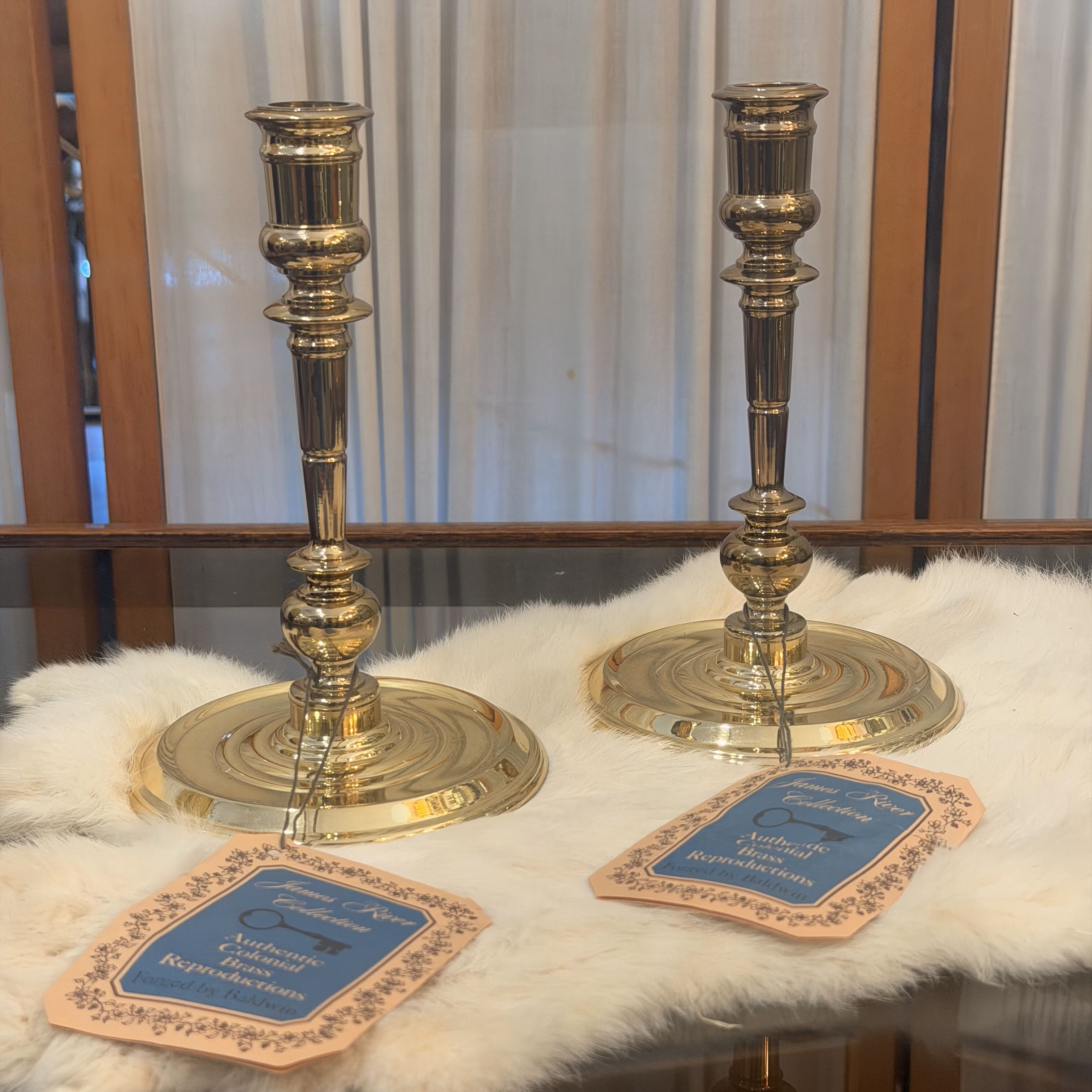 A pair of ornate brass candlesticks with circular bases, displayed on a white faux fur rug with two blue and gold cards in front of them.