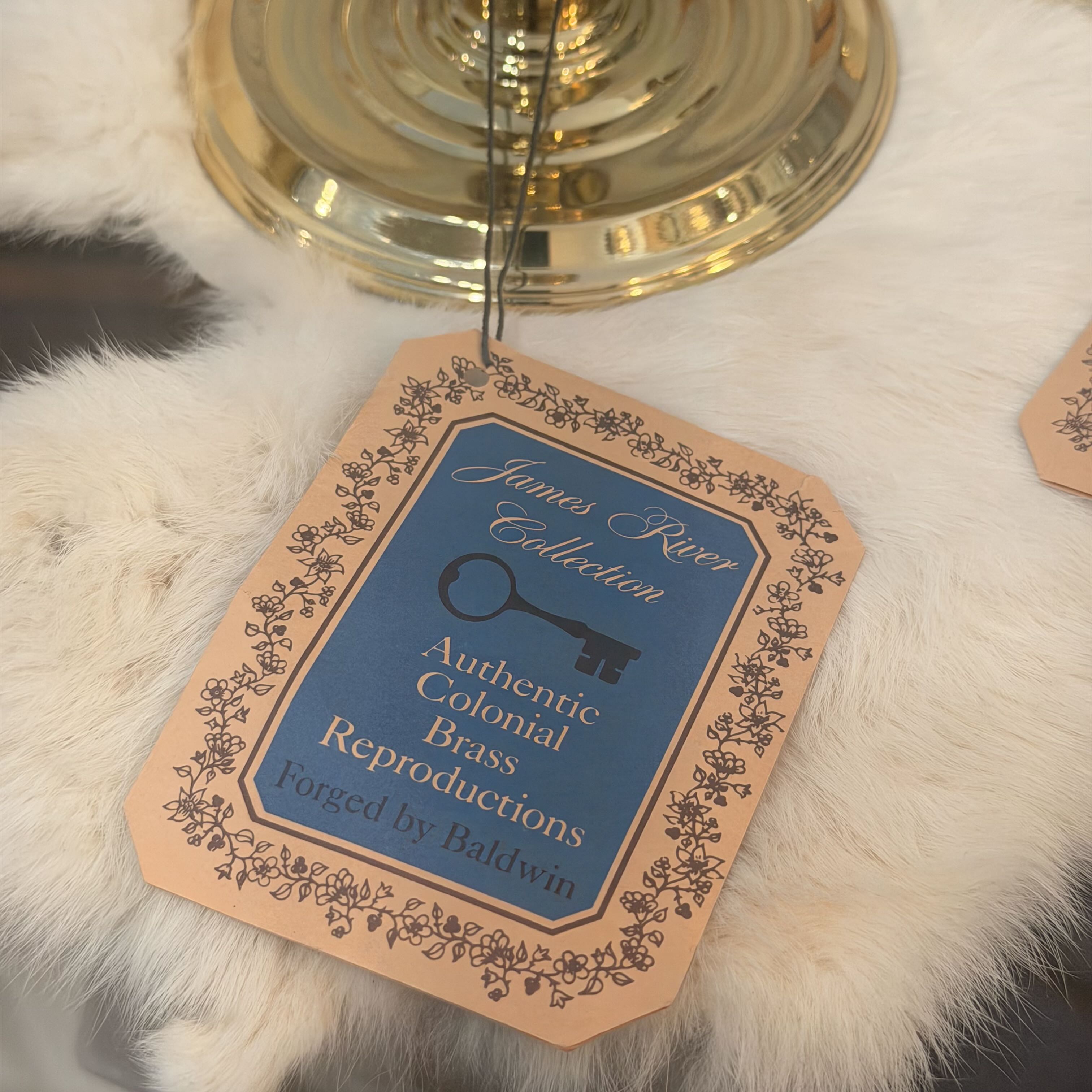 A hanging paper tag for the James River Collection Authentic Colonial Brass Reproductions, styled on a white faux fur surface with a gold brass candle holder base in the background.