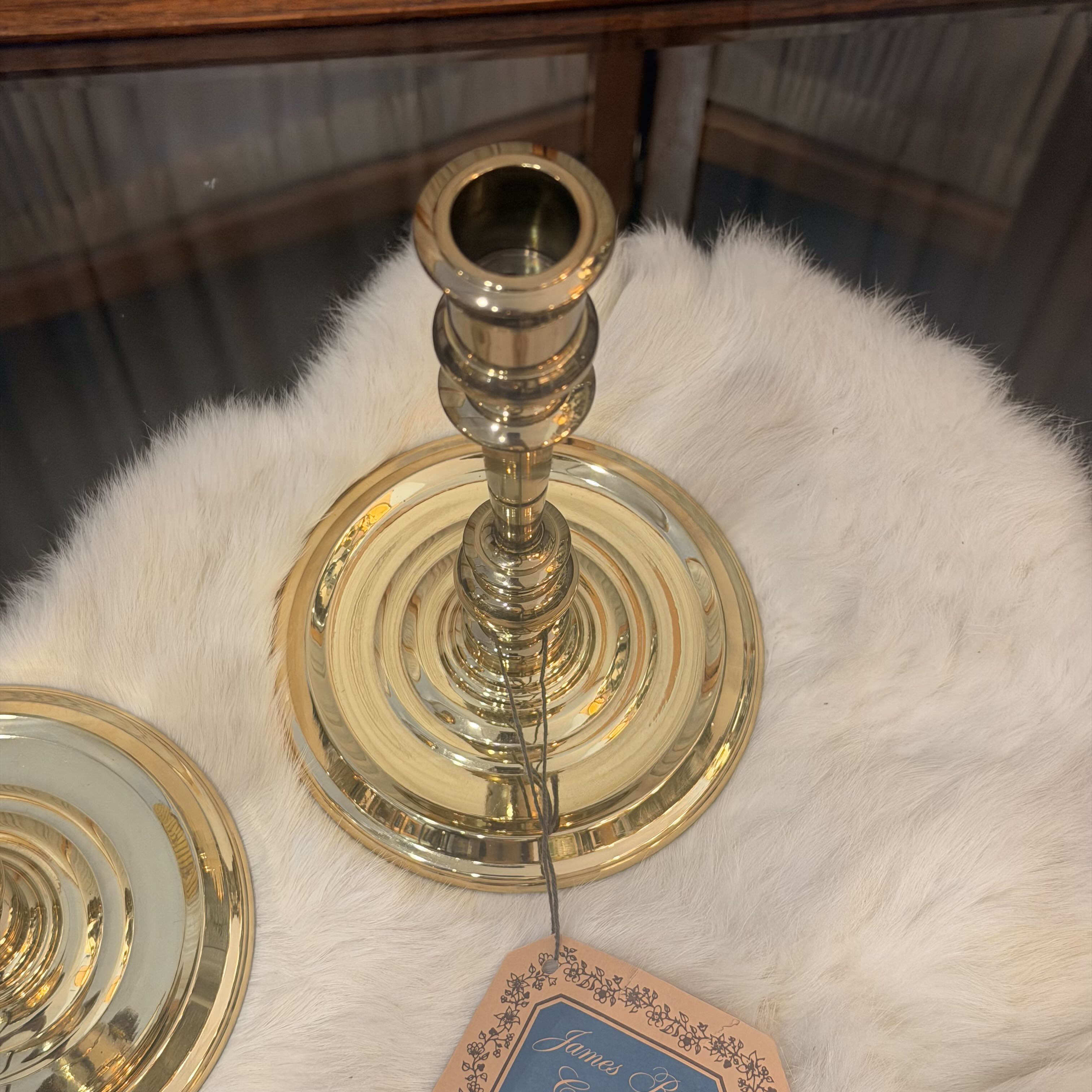 A polished brass candlestick holder with a circular base and tiered stem, displayed on a white faux fur surface with part of another matching piece visible at the lower left.