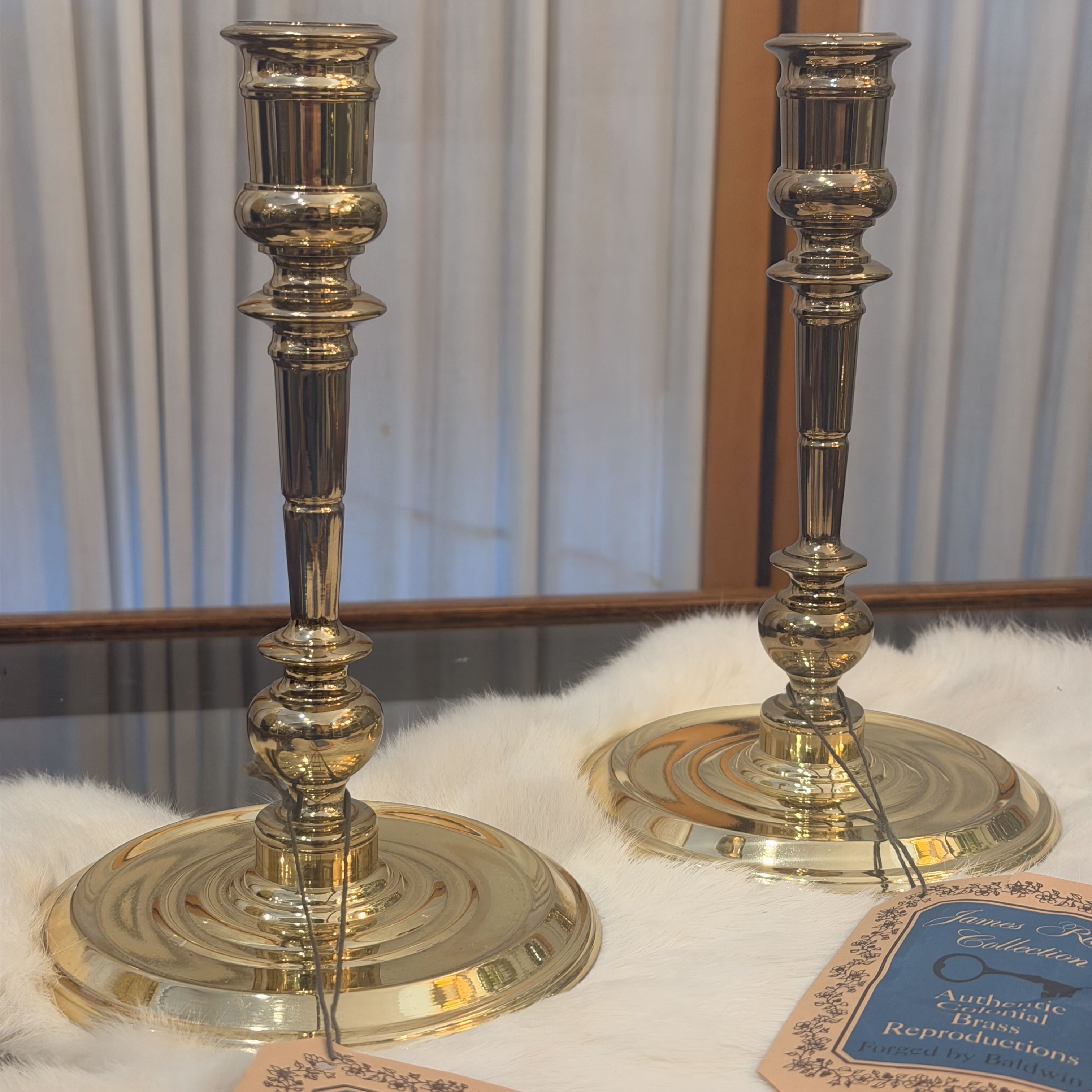 A pair of polished brass candlesticks with ornate fluted stems and round tiered bases, displayed on a white faux fur surface with a glass tabletop and curtain background.