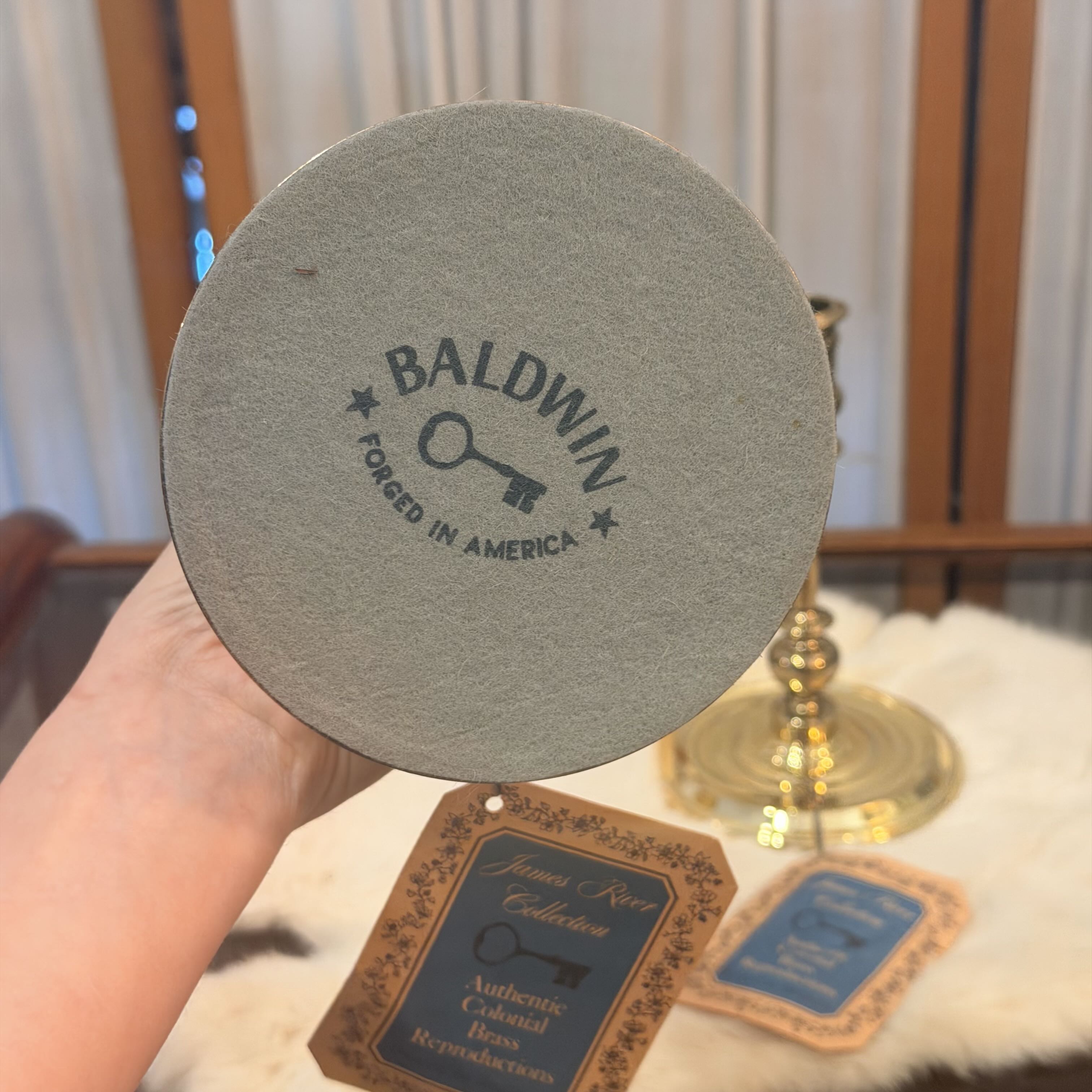 A hand holds a round gray coaster printed with the words 'BALDWIN FOCUSED IN AMERICA' and a key graphic. A hanging tag below reads 'James J.