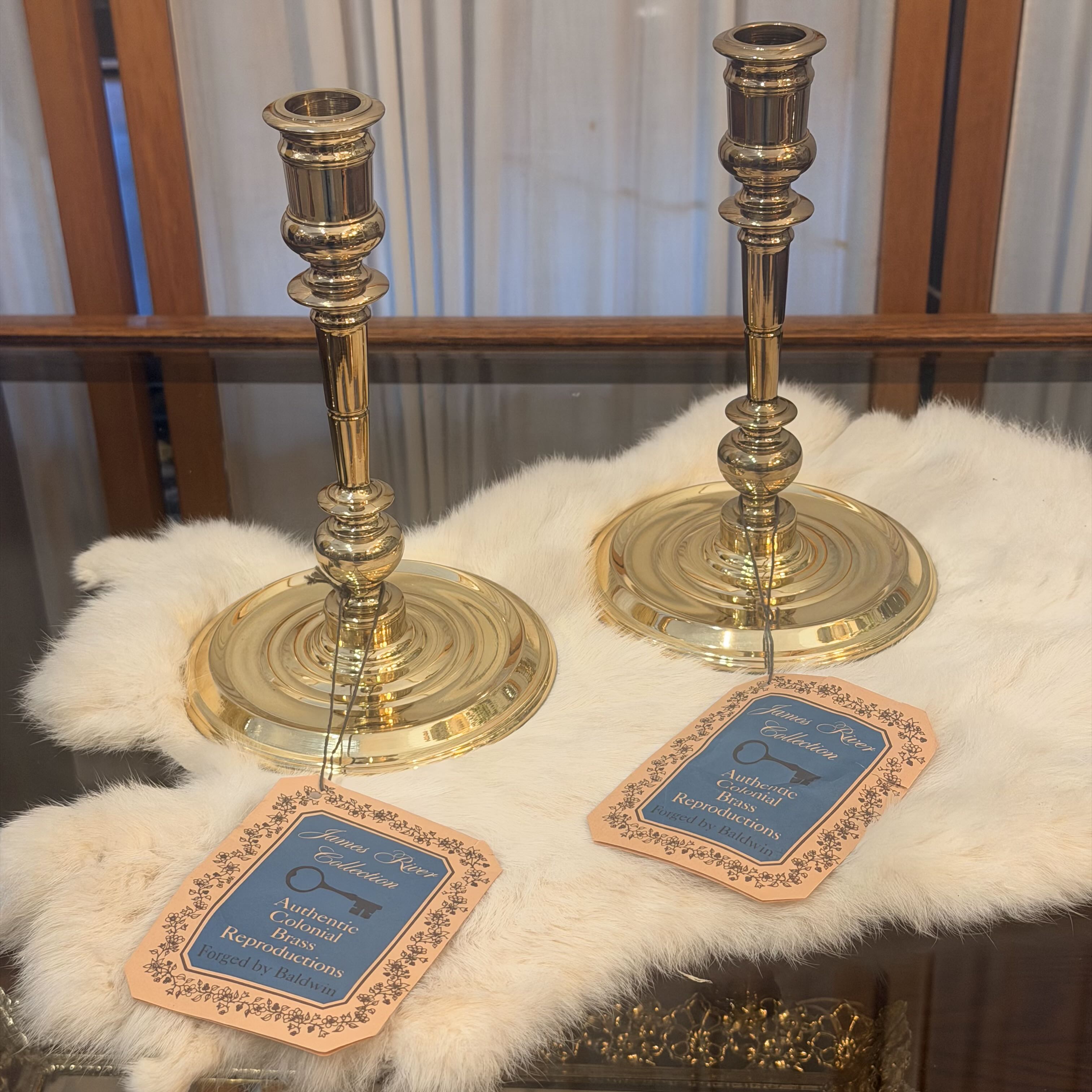Two ornate brass candlesticks displayed on a white faux fur surface with decorative tags in front, set against a background of wooden furniture and curtains.