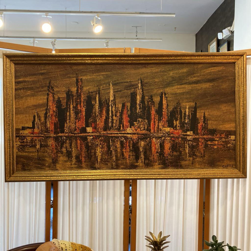 A large framed painting depicting a dark, textured city skyline with tall spires and reflections in the water, housed in an ornate gold frame. The painting is displayed hanging on a white curtain wall in an indoor showroom setting.
