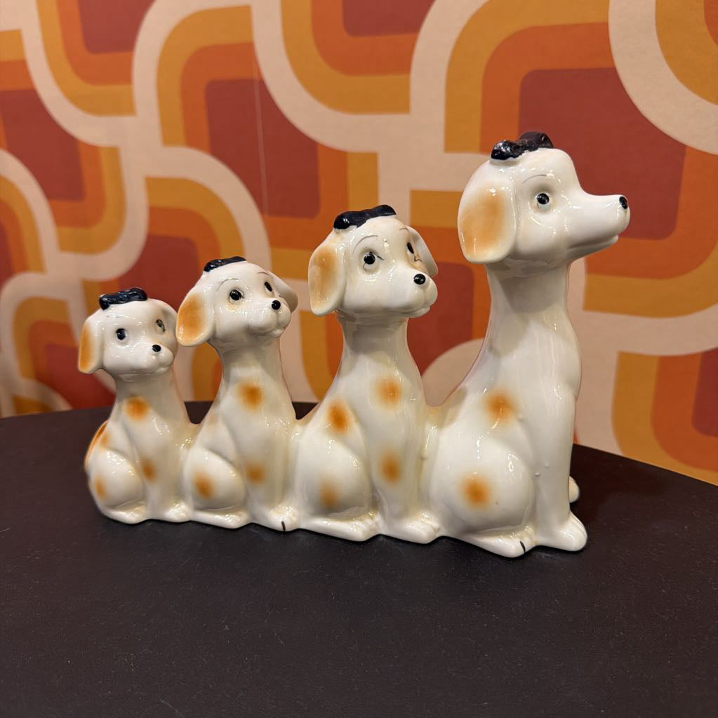 Three ceramic Dalmatian dog figurines in a row, decorated with orange spots and black ears, sitting on a dark surface in front of a retro orange and white patterned background.
