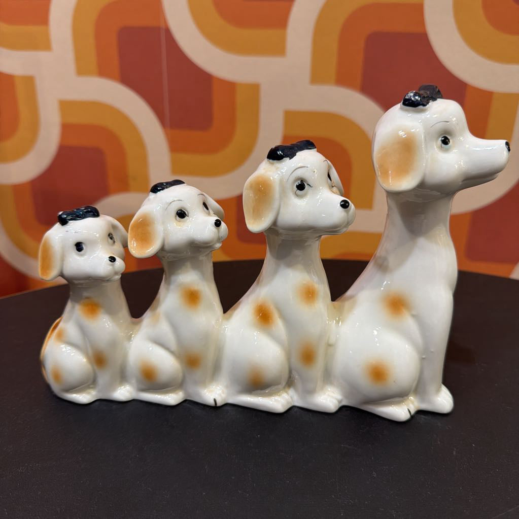 Three ceramic Dalmatian dog figurines in white with orange spots and black noses, arranged in a row on a dark surface against a retro orange and cream patterned background.
