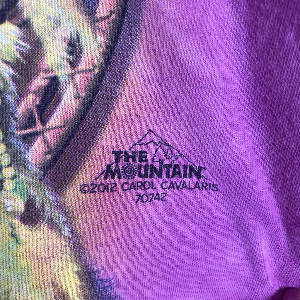 Close-up of a purple graphic t-shirt featuring a black printed logo that reads 'THE MOUNTAIN' with a mountain graphic, 'C©2012 CAROL CAVALARIS', and '70742'.