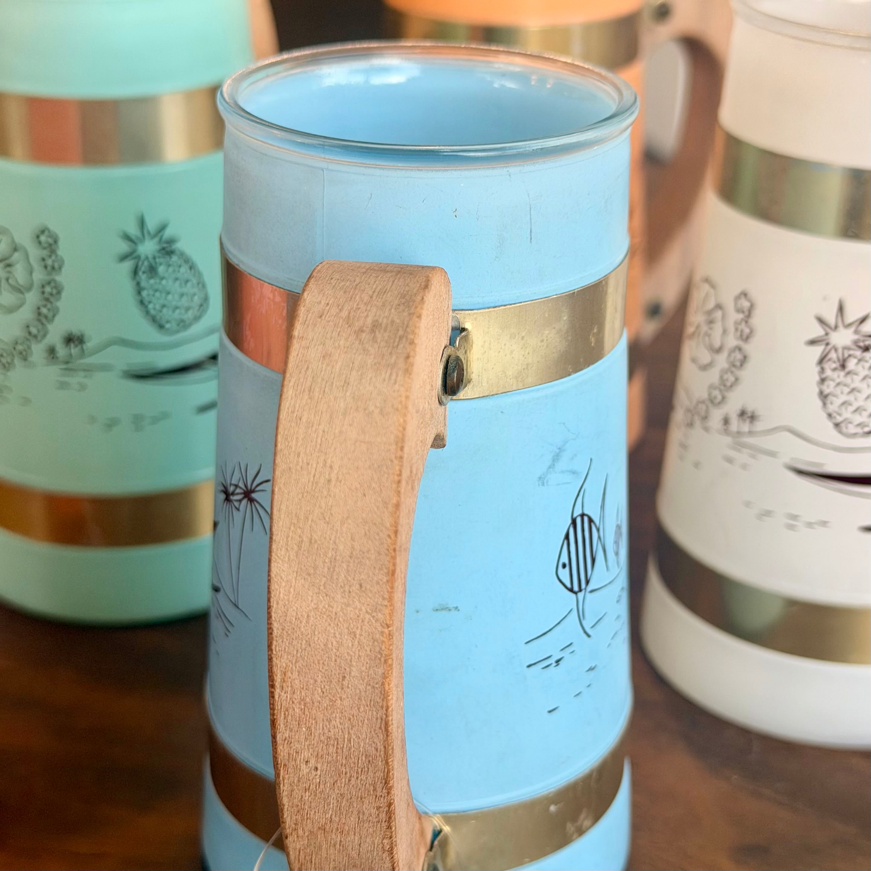 A light blue ceramic mug with a wooden handle, gold metal bands, and black tropical line drawings, shown on a wooden table with other mugs in the background.