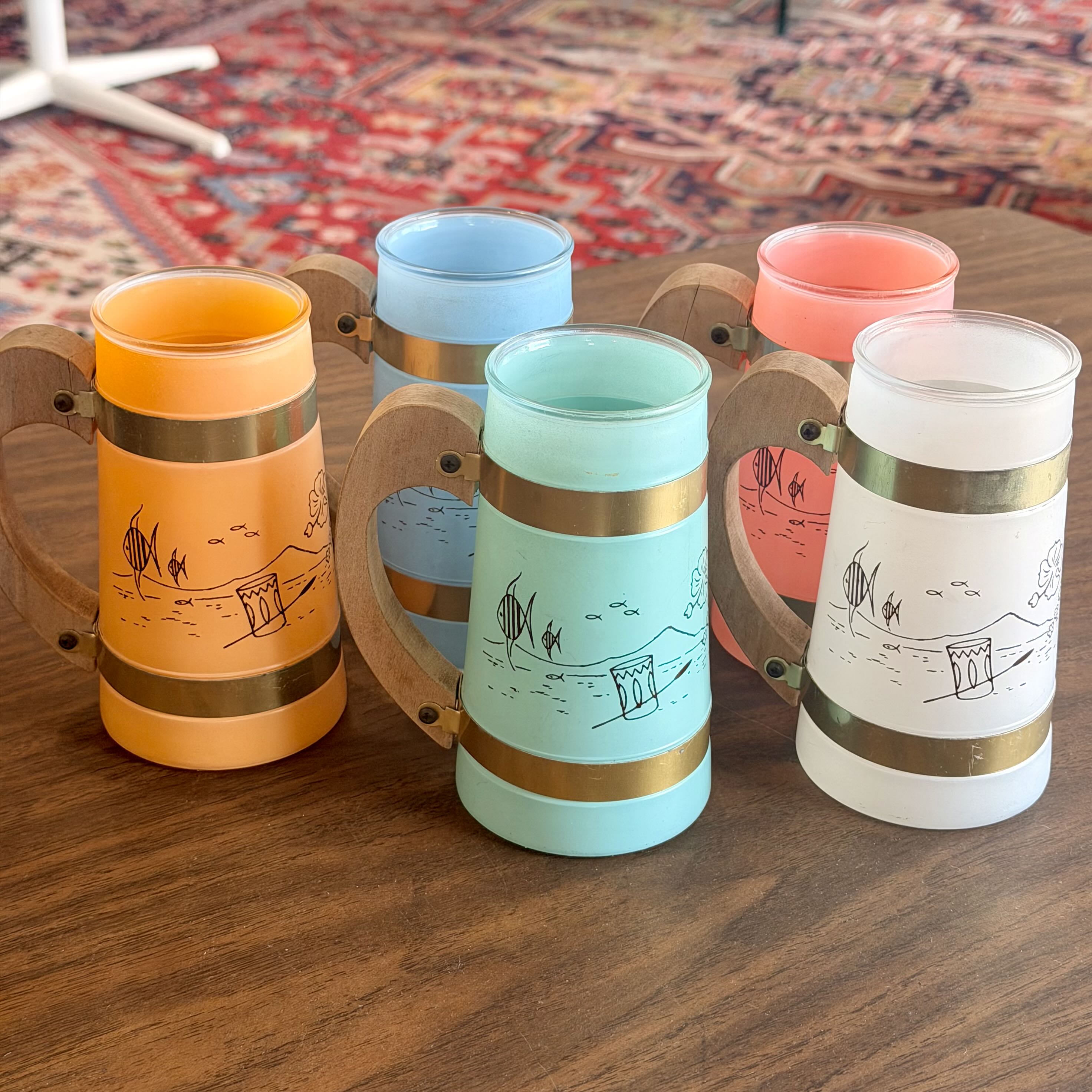 Four frosted glass beer steins with wooden handles and gold bands, displayed on a wooden table. The steins are in orange, light blue, coral pink, and white, each decorated with a black line-art illustration of a beach and a beer mug.