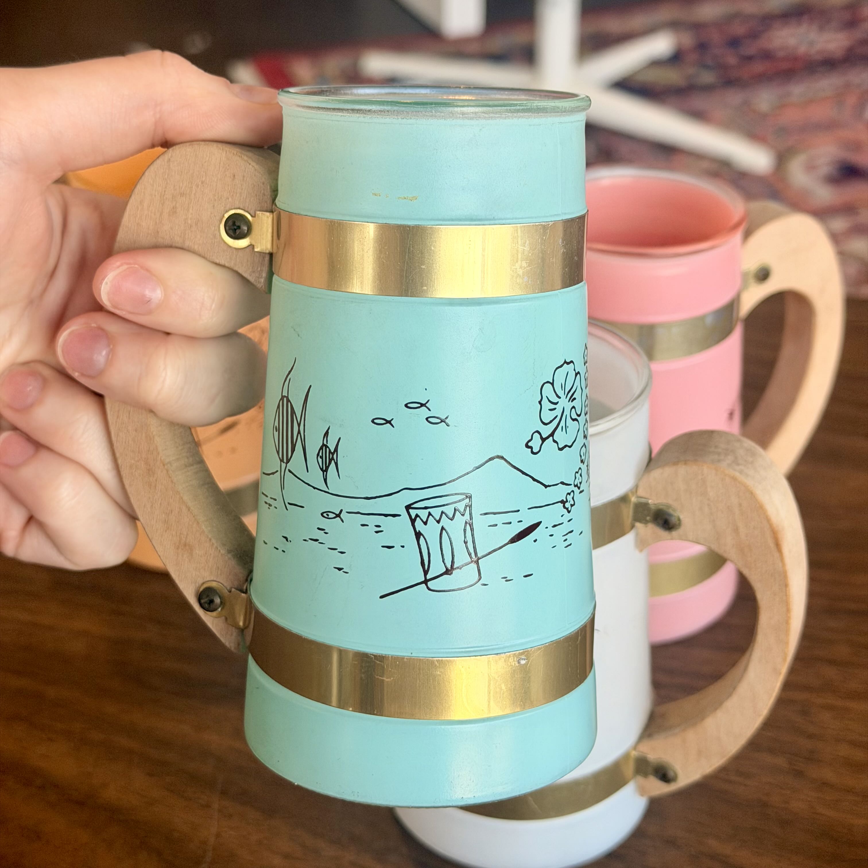 A hand holds a frosted light blue glass mug with a wooden handle and gold metal bands, decorated with a simple black line drawing of a beach scene. A pink and a white version of the same mug are partially visible in the background on a wooden table.