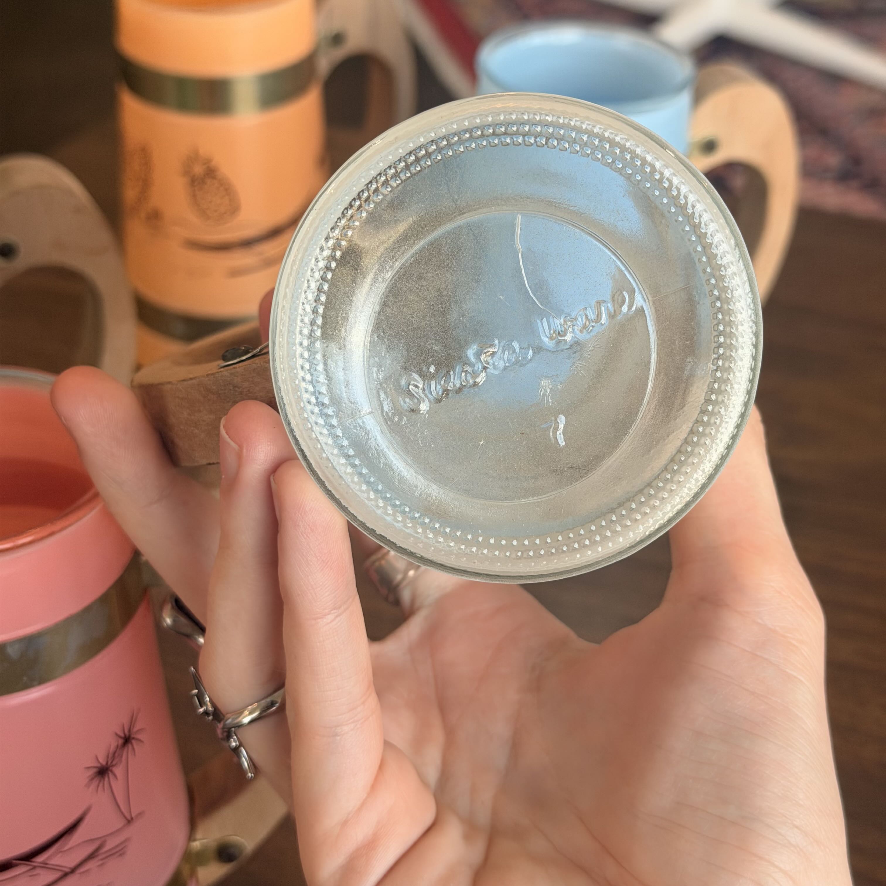 A hand holding a clear glass cup with a beaded rim and embossed brand text inside, with other colorful cups in the blurred background.