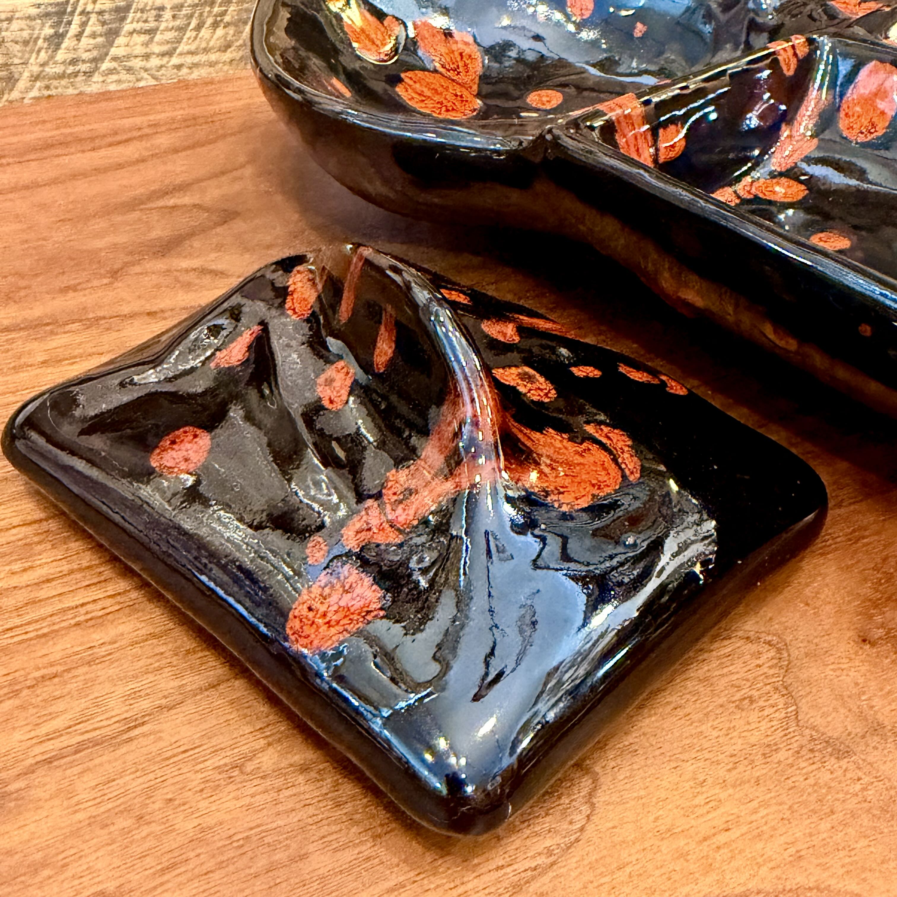 A glossy black ceramic dish with orange speckled patterns, shown on a wooden surface next to another matching dish.