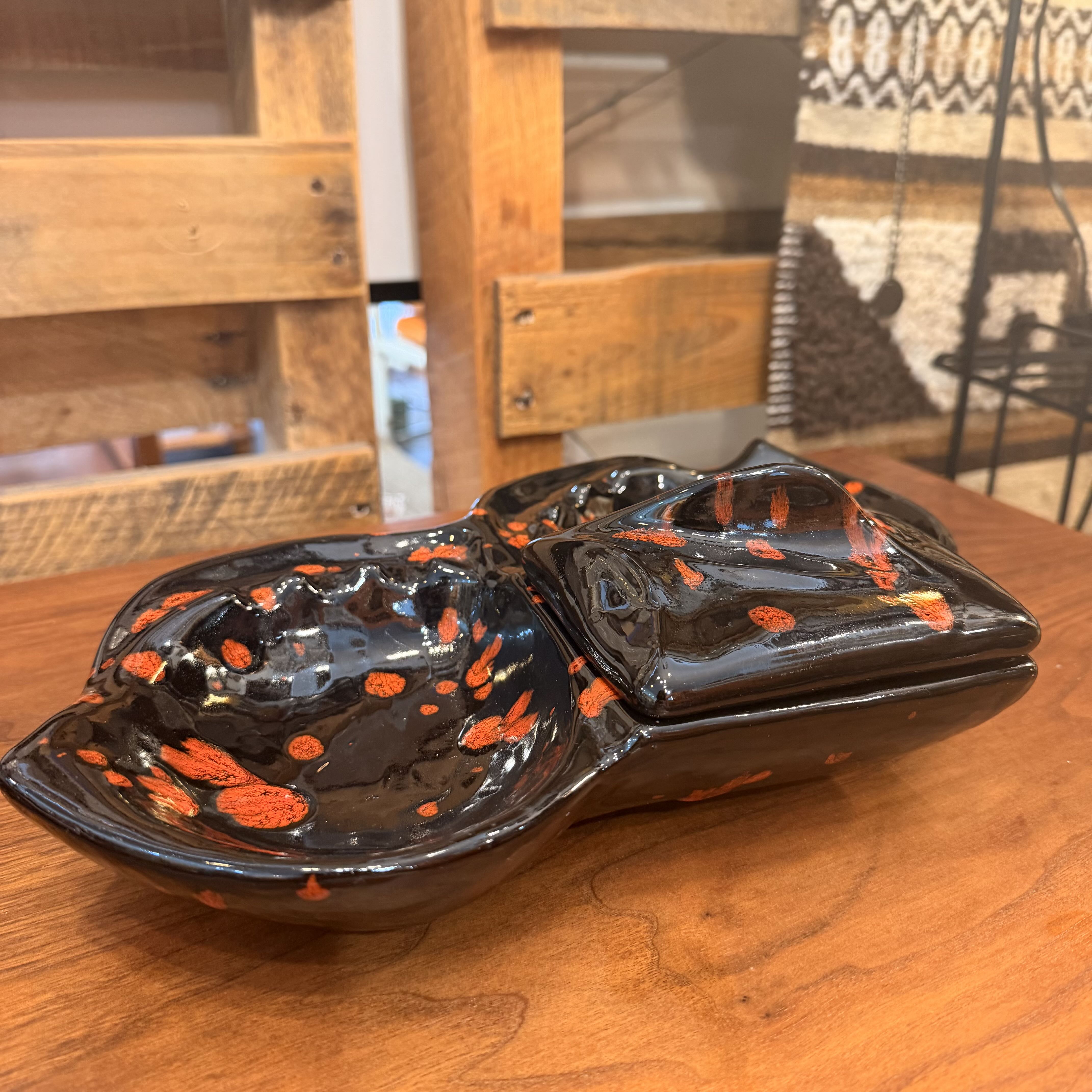 A glossy black ceramic dish with a matching lid, decorated with bright orange speckles, displayed on a wooden table in a rustic indoor setting.