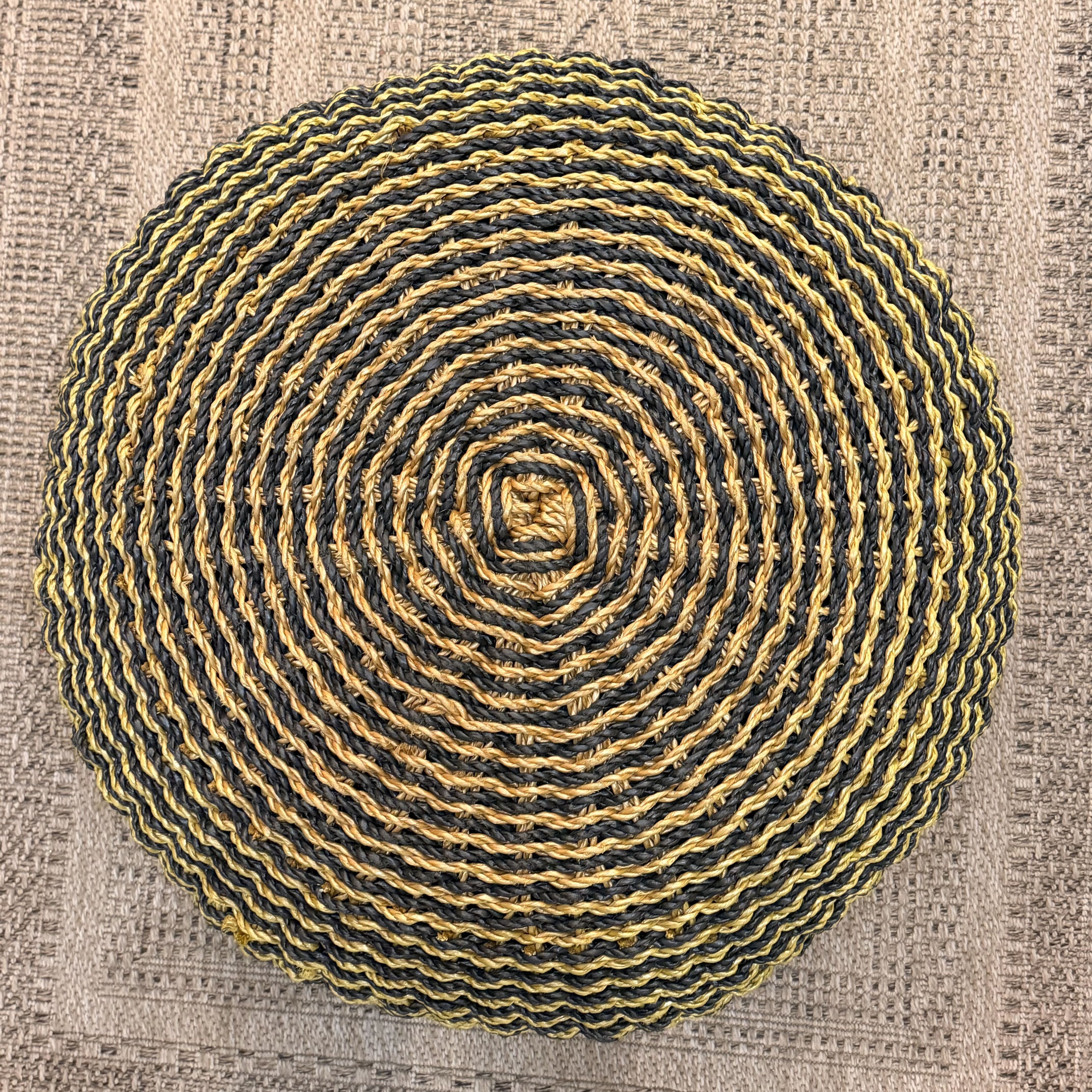 A round woven pouf cushion with concentric rings of yellow and black yarn, photographed from directly above on a textured beige fabric background.