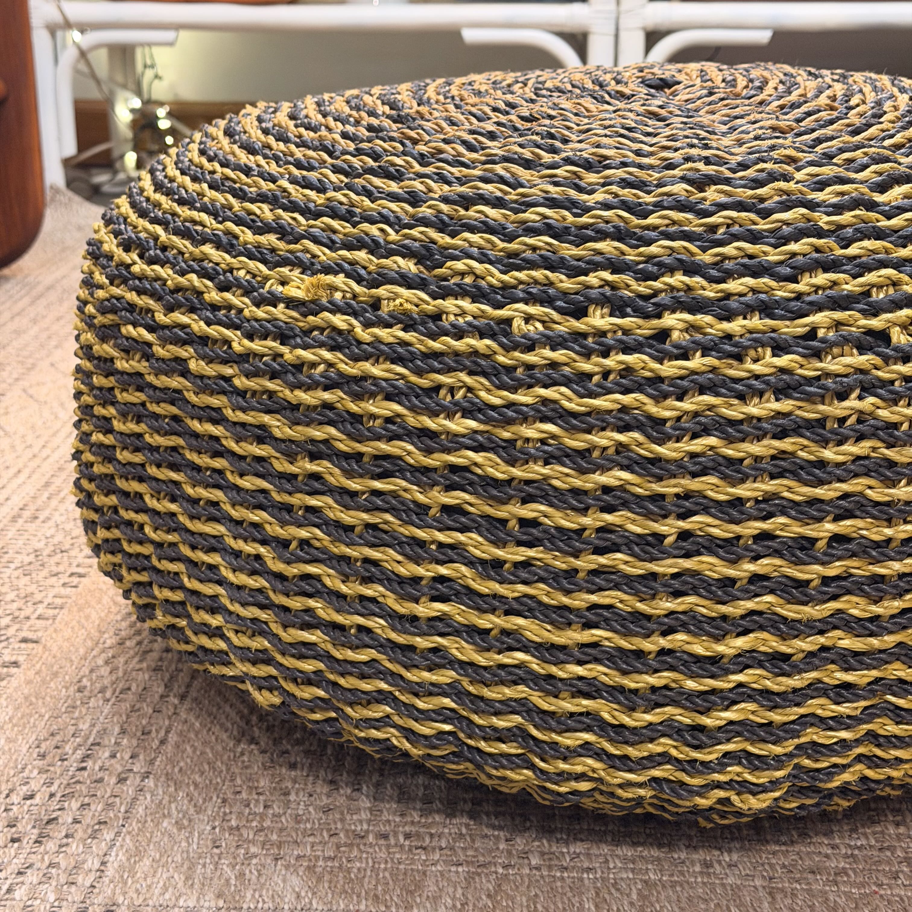 A round woven pouf ottoman with a textured pattern of dark gray and mustard yellow horizontal stripes, resting on a woven rug.