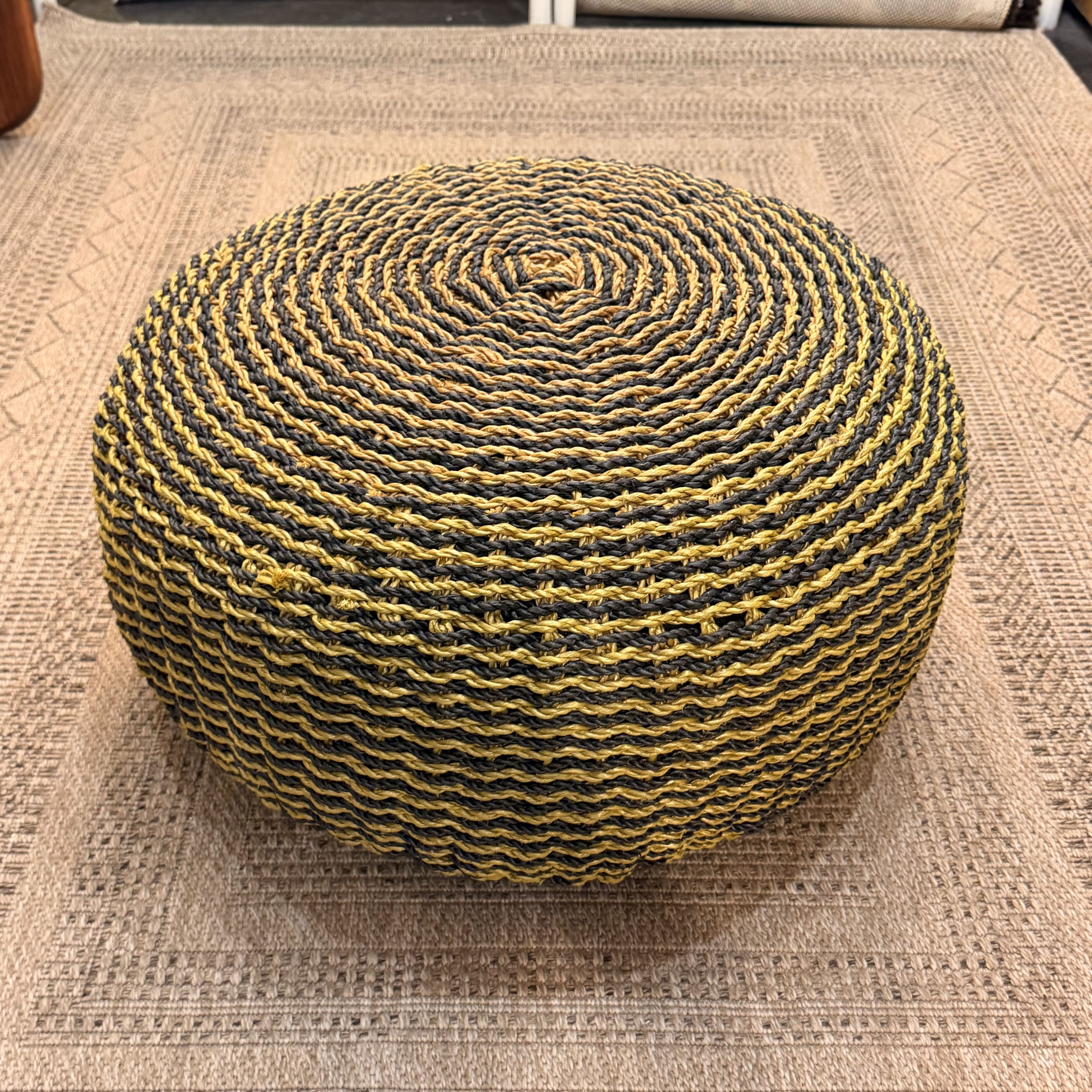 A round woven pouf ottoman with a spiral pattern of black and mustard yellow rope, placed on a textured beige woven rug.