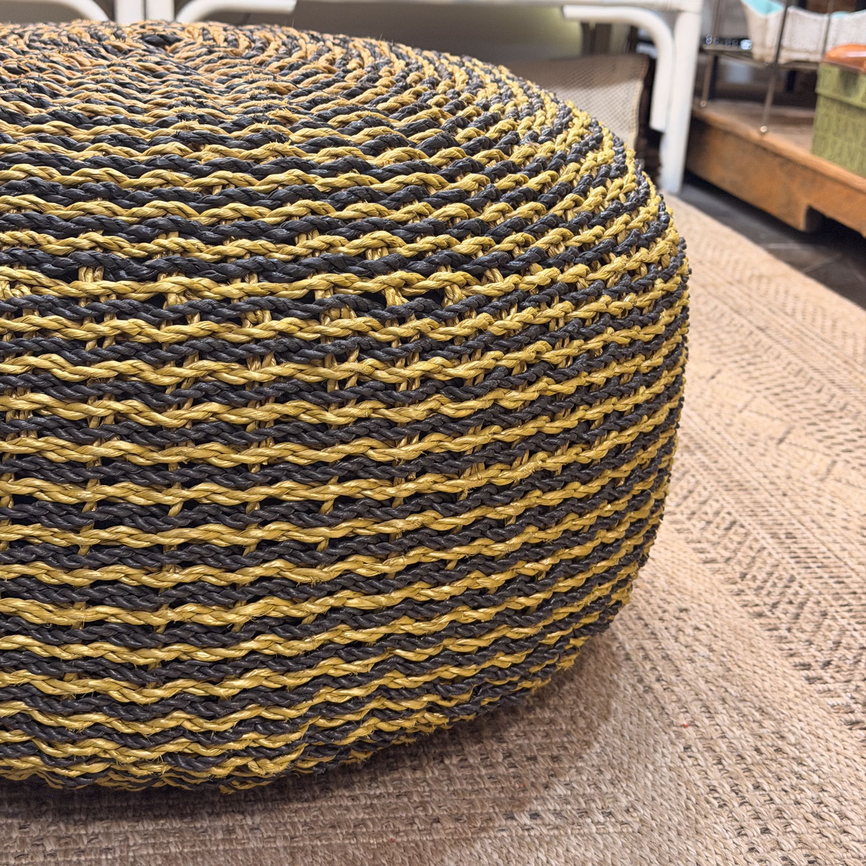 A close-up of a round woven pouf ottoman with alternating dark gray and mustard yellow horizontal stripes, resting on a textured beige rug.