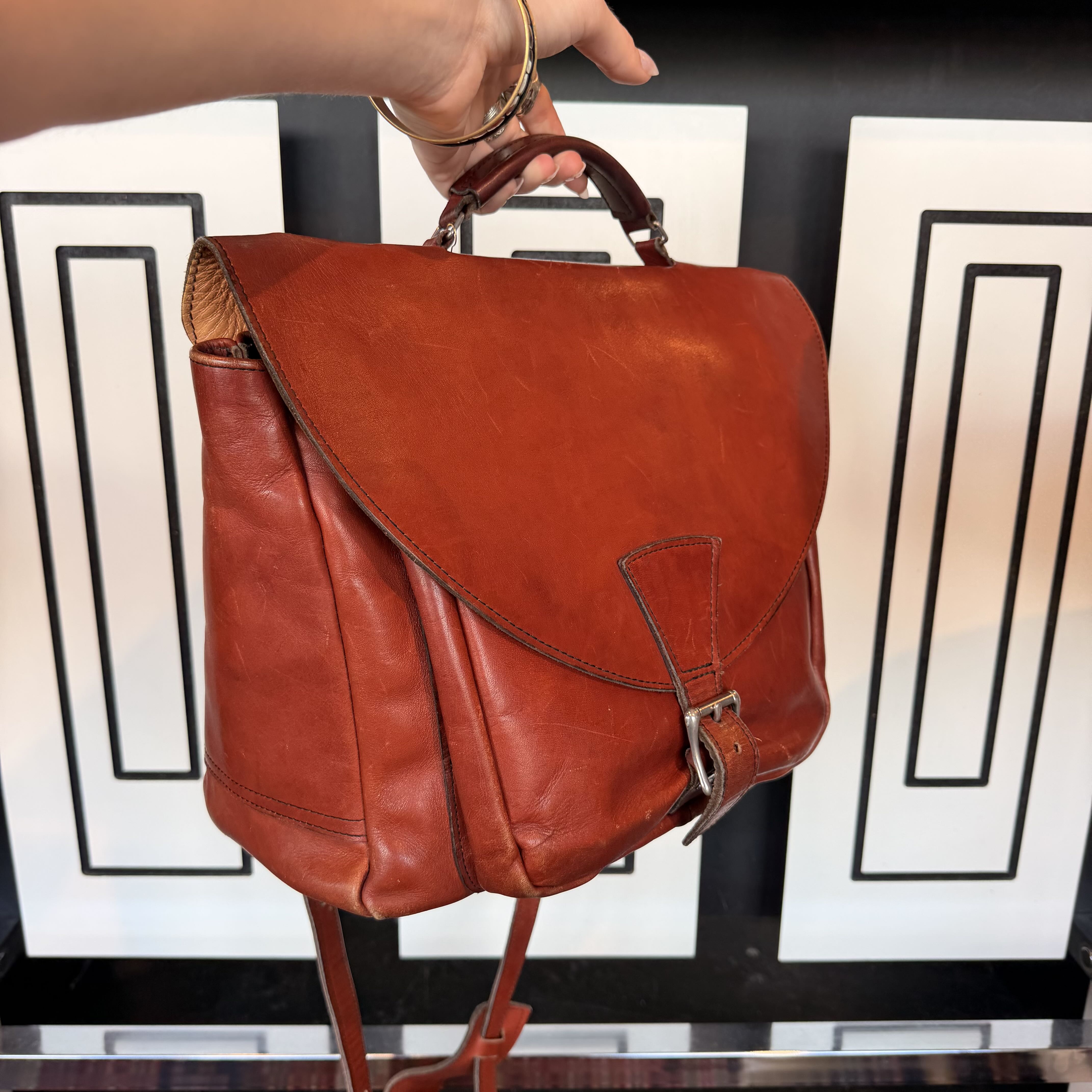 A reddish-brown leather backpack with a top handle and buckle closure, held by a hand in front of a black and white geometric background.
