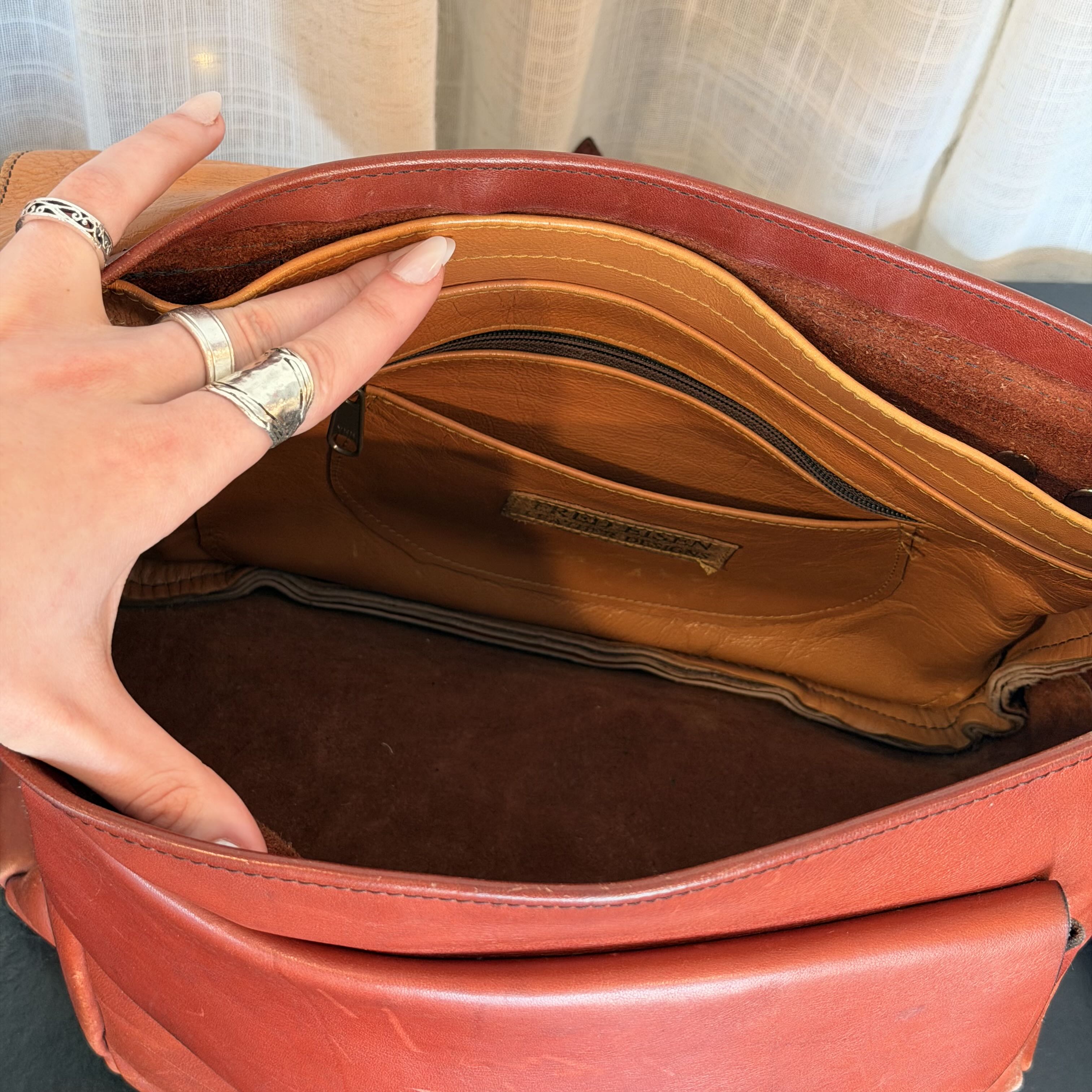 Close-up of a person's hand inside an open reddish-brown leather bag, showing the tan interior lining, card slots, and dark brown suede-like bottom compartment.