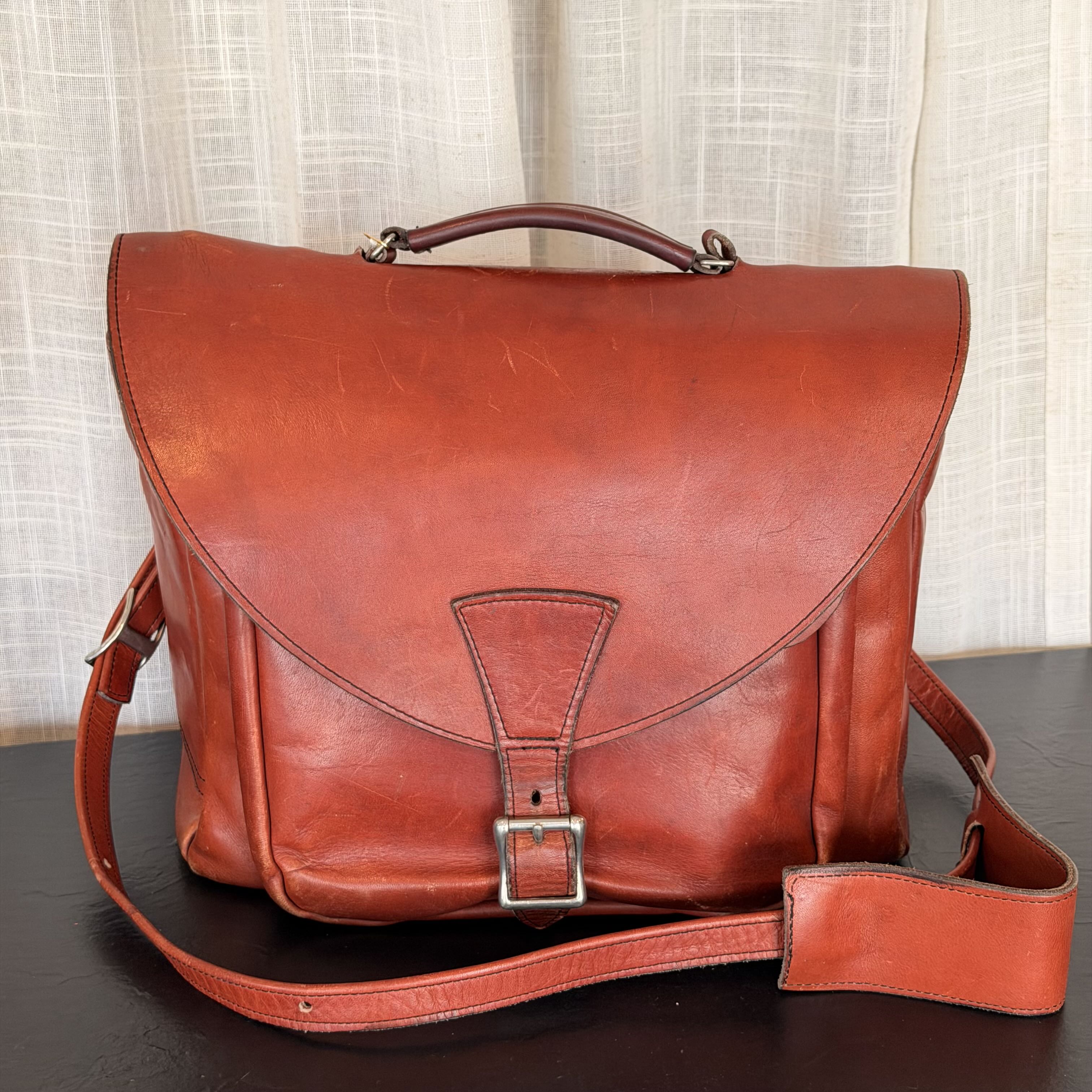 A reddish-brown leather messenger bag with a top handle, adjustable shoulder strap, and front buckle closure, displayed on a dark surface against a light curtain background.