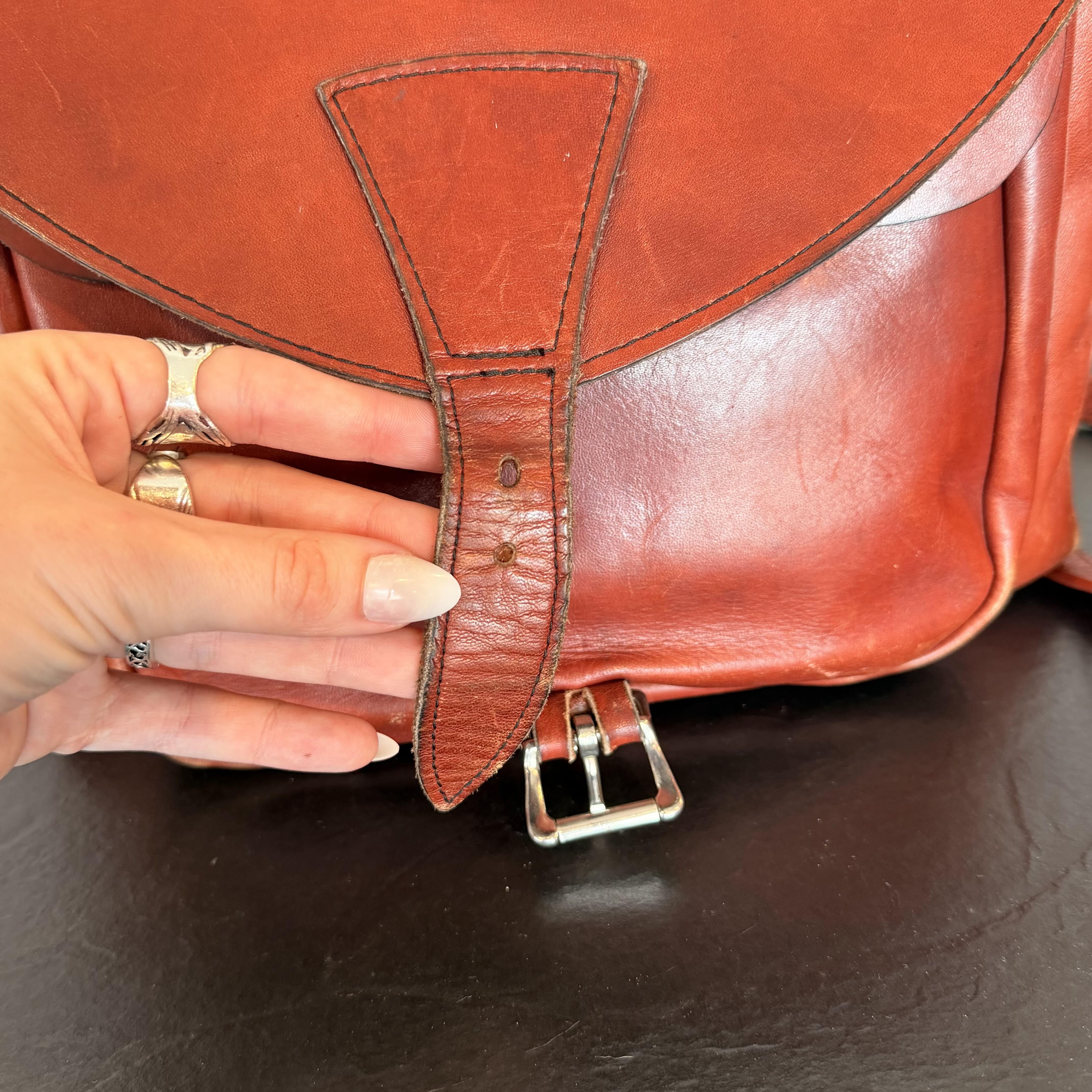 A close-up of a reddish-brown leather satchel or backpack being held by a hand, showing the front flap tab and metal buckle on a dark surface.