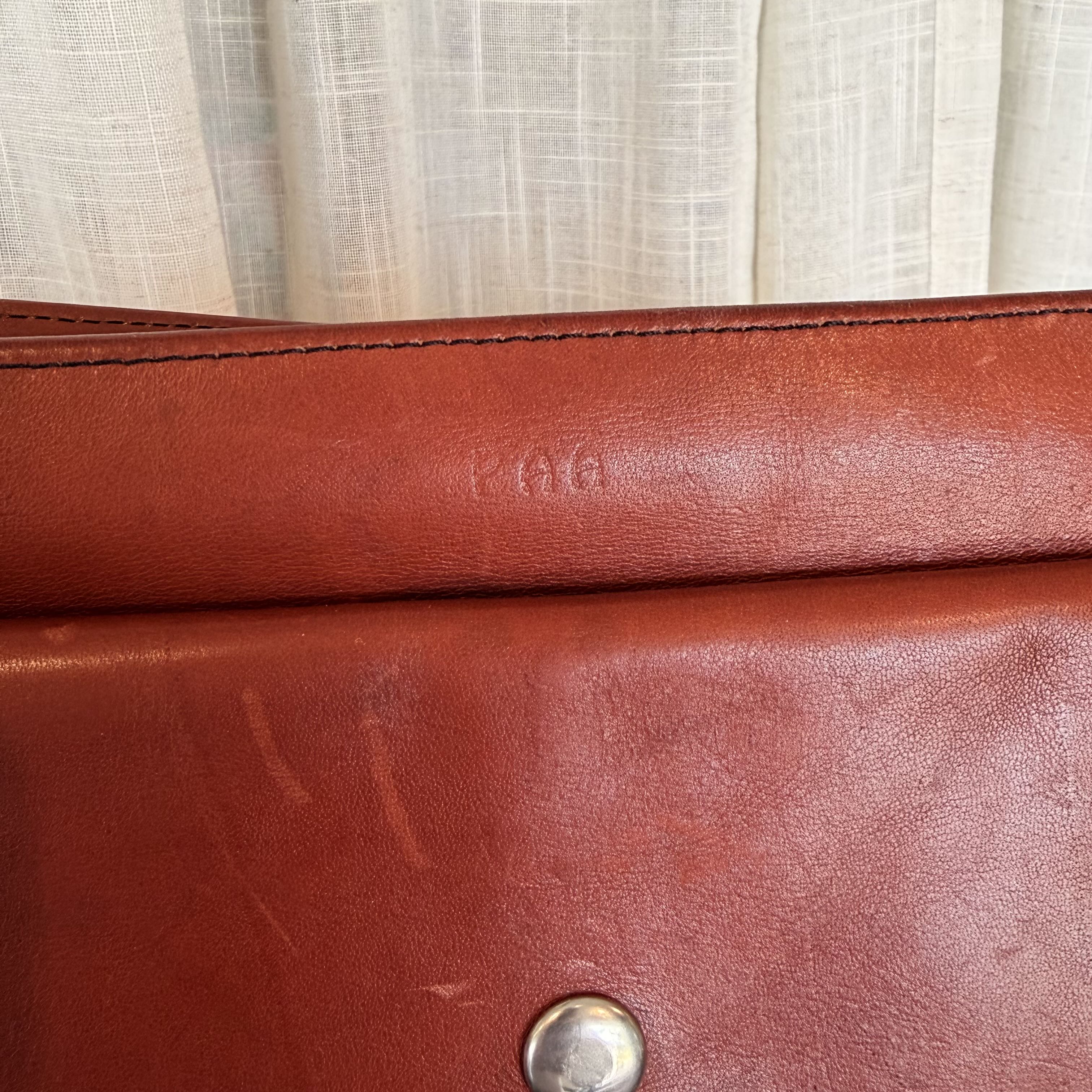 Close-up of a reddish-brown leather bag or wallet showing the embossed brand name 'KAE' and a silver snap button, with a cream curtain background.