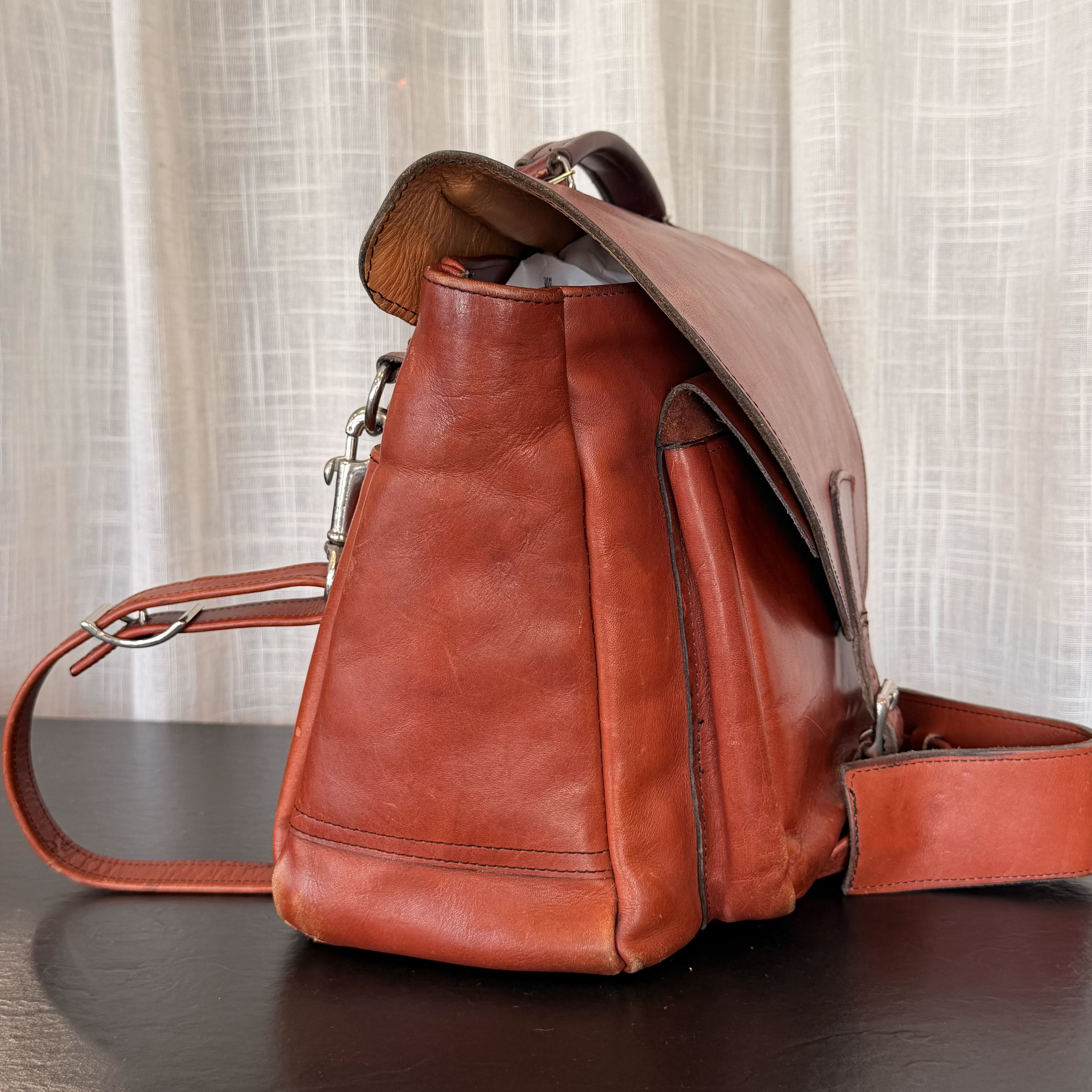 A reddish-brown leather backpack with a flap closure and shoulder strap, resting on a dark wooden surface in front of a light beige curtain backdrop.