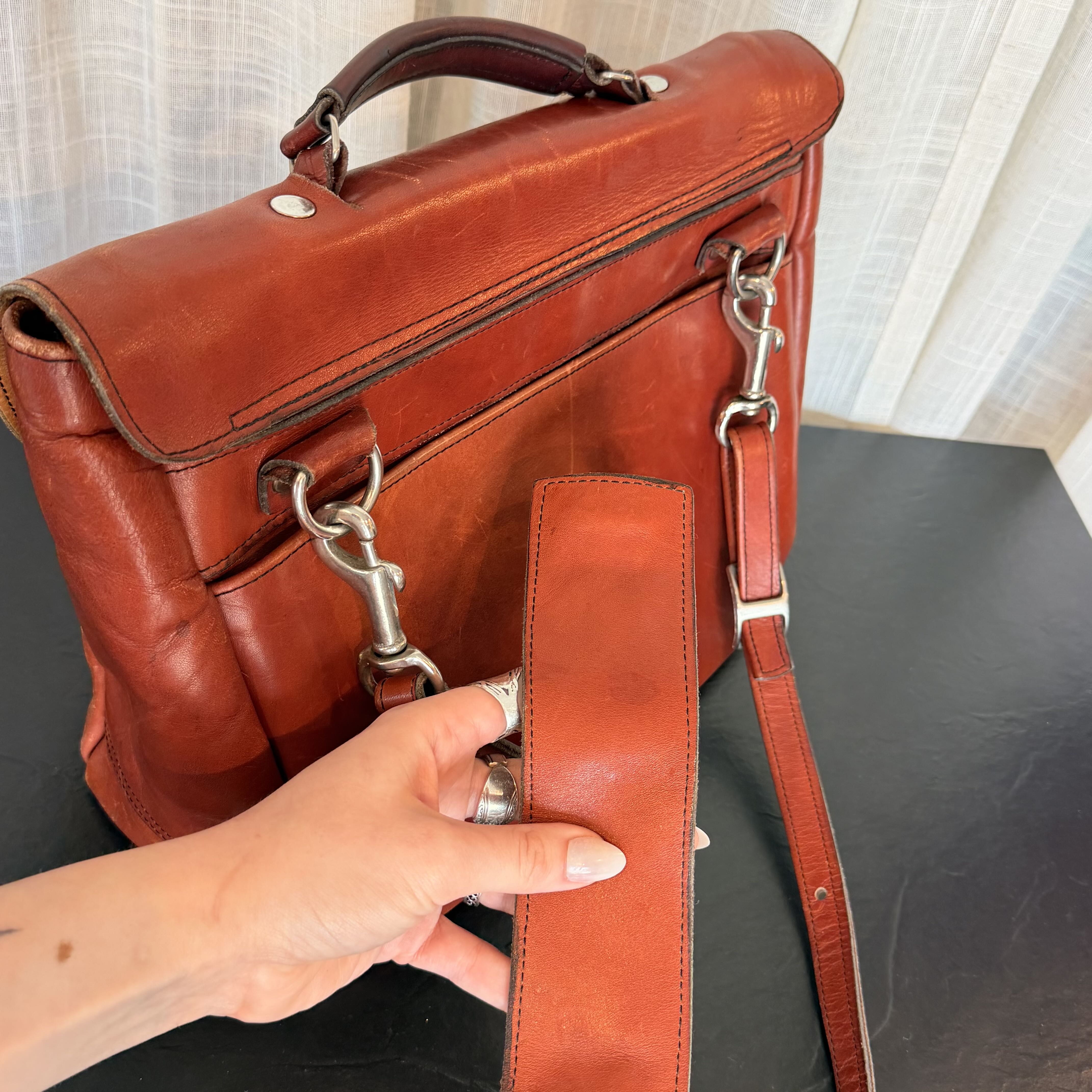 A close-up of a person's hand holding a matching leather strap in front of a rust-brown leather briefcase or satchel with silver hardware, set on a dark tabletop with a curtain background.
